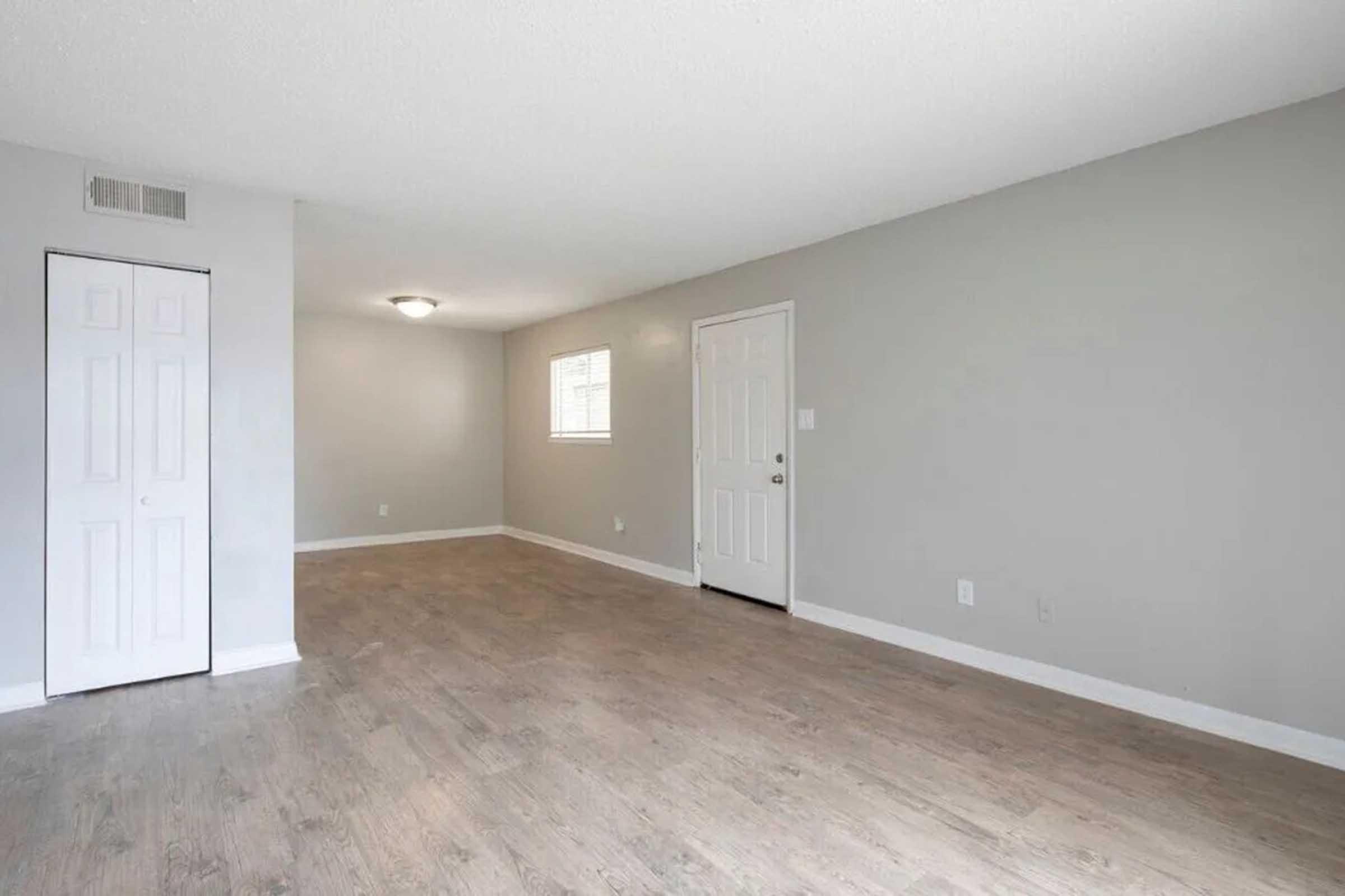 A spacious, empty room with light gray walls and a light wood floor. There is a small window allowing natural light, a ceiling light fixture, and two doors: one leading to a closet and the other to an entryway. The room has a clean, minimalist aesthetic.