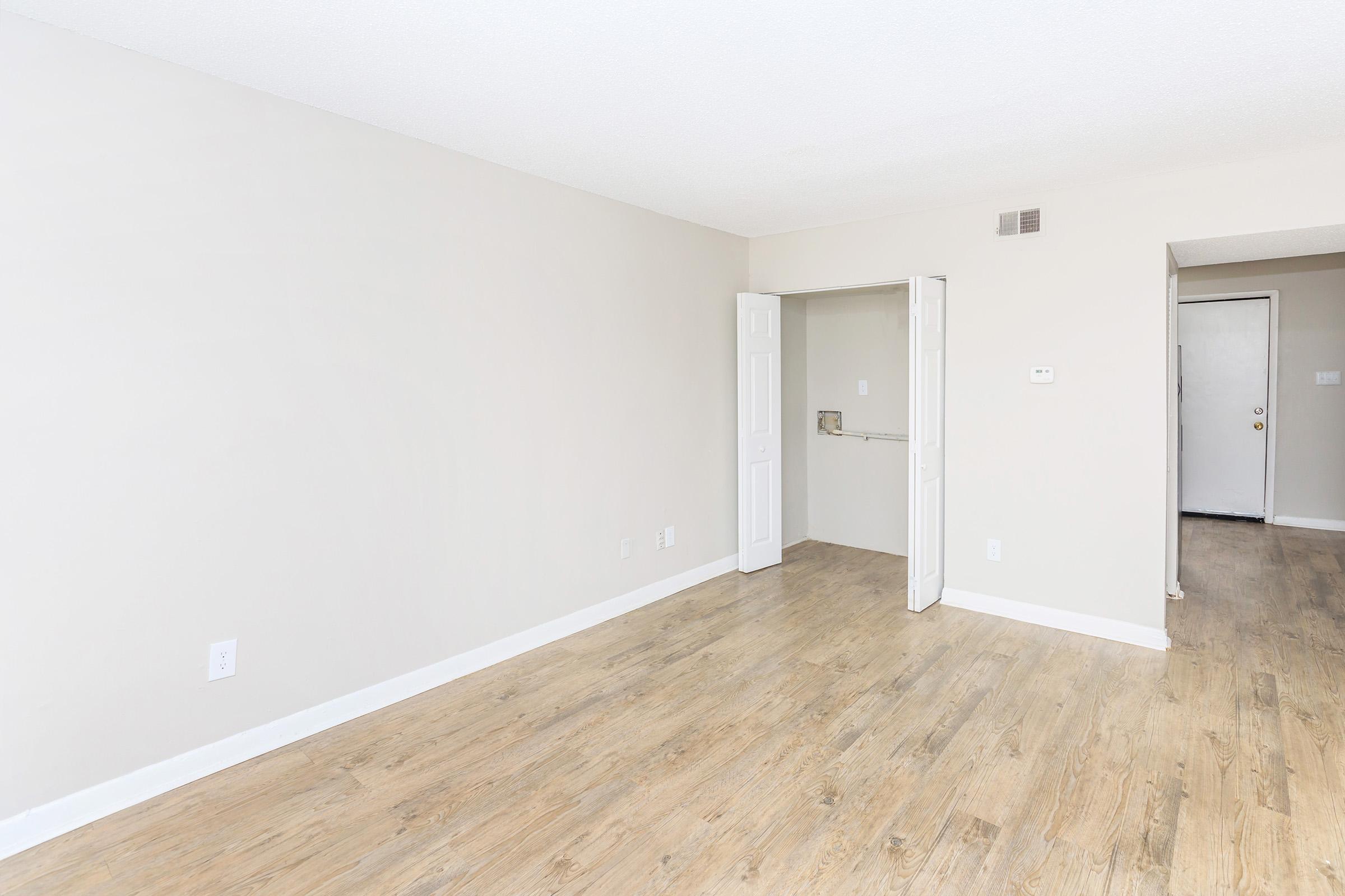 A spacious, empty room with light-colored walls and a wood-like floor. There is an open closet with a door on the left side and a doorway leading to another area on the right. The room is well-lit, creating a bright and airy atmosphere.