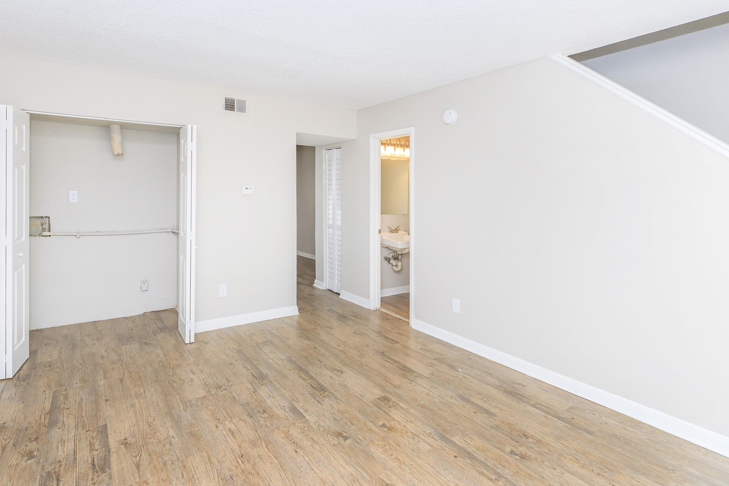 An empty room with light-colored walls and a wooden floor. There are two open doors leading to other spaces; one appears to lead to a bathroom and the other to a hallway. Natural light enters the room from a nearby window, enhancing the airy feel of the space.
