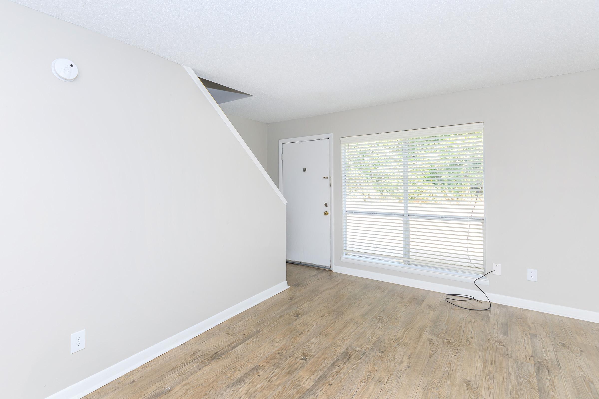 Interior of a light-filled room featuring a white wall, a staircase leading to an upper level, a door with a window next to it, and hardwood flooring. Sunlight is streaming through the window, highlighting the space's neutral tones and minimalist design.