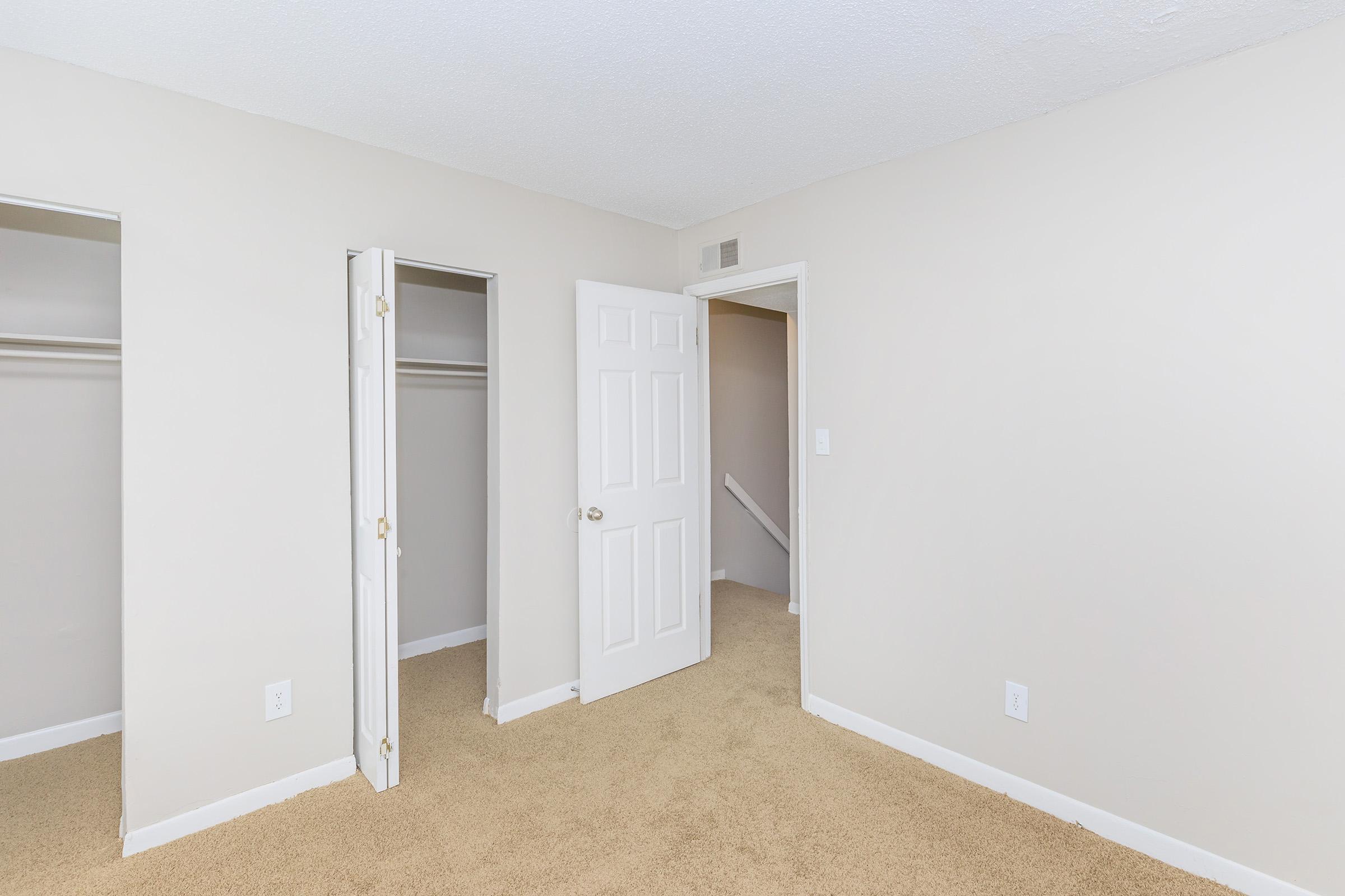 Empty bedroom with beige walls and tan carpeting. Two closet doors on the left, an open door leading to another room on the right, and a staircase visible in the background. Lighting is bright, creating a spacious and neutral atmosphere.