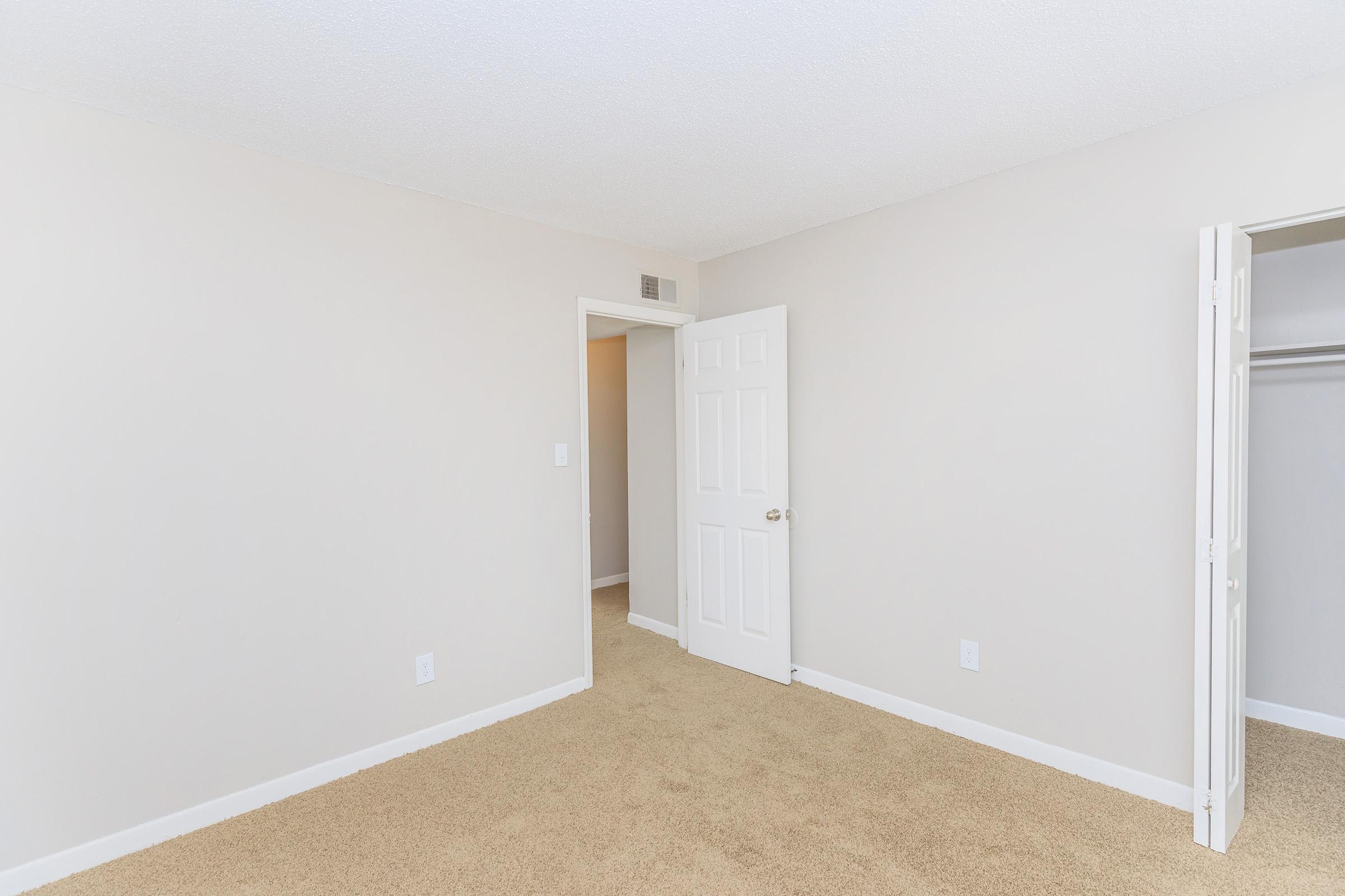A clean, empty room with beige carpet and light-colored walls. A white door is partially open, leading to another space, while a closet door is also ajar, showcasing the interior. The room has a bright, neutral atmosphere, suitable for various uses.