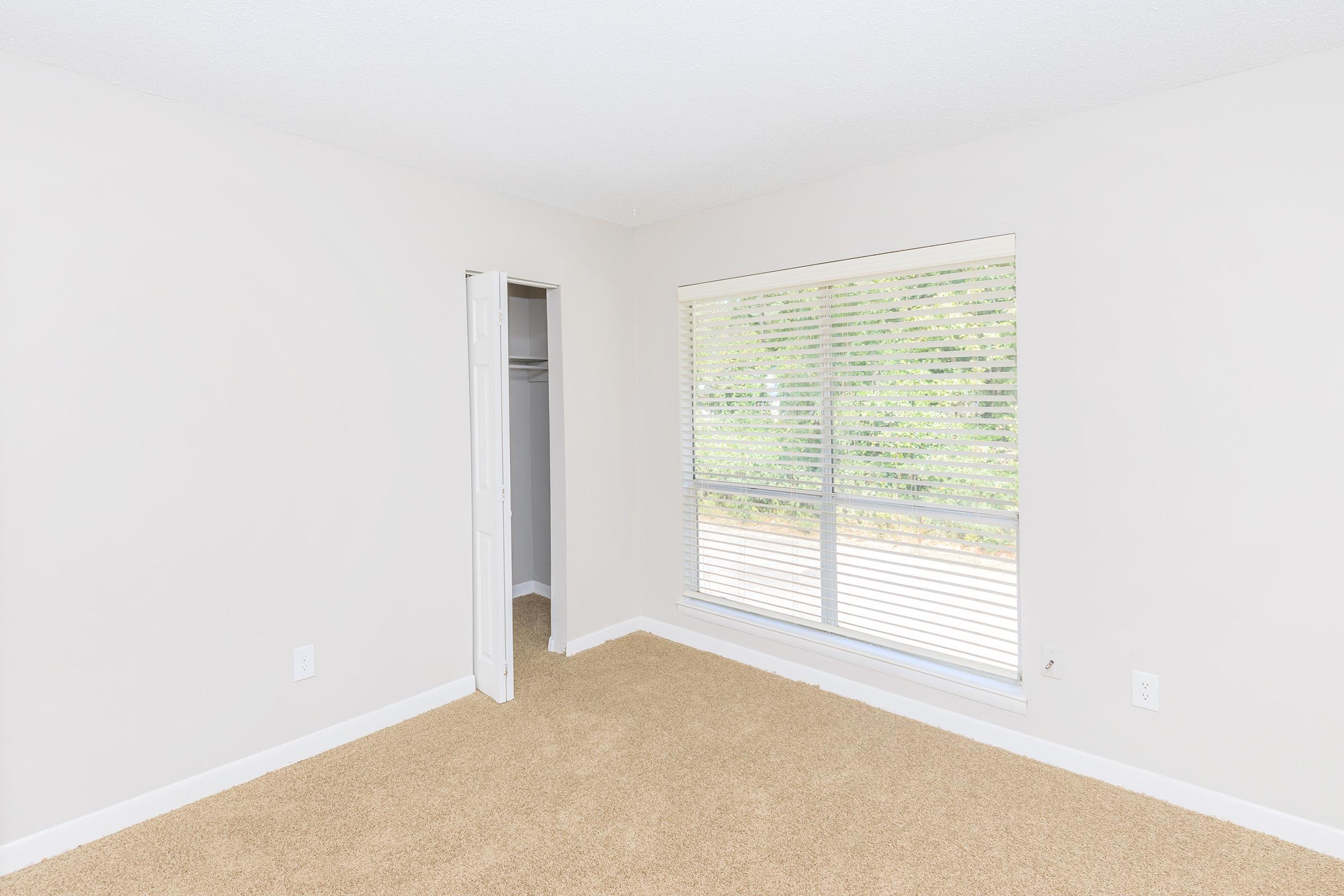 A spacious, empty bedroom featuring beige carpet flooring and light-colored walls. There is a large window with blinds that lets in natural light, and a closet with double doors visible to the left. The overall atmosphere is bright and simple, ideal for personalized decor.