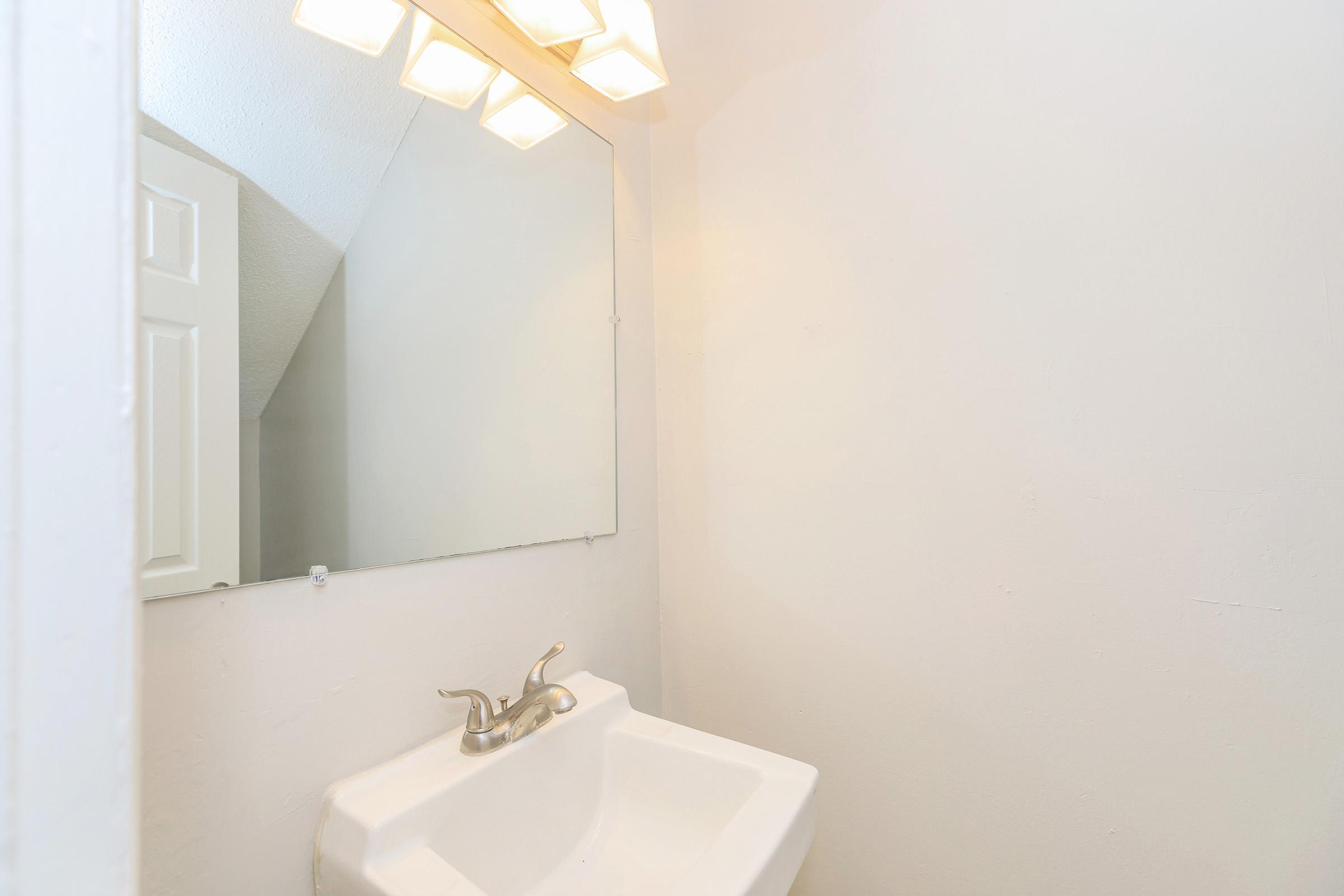 A clean, minimalist bathroom featuring a white sink and a large mirror above it. The walls are painted white, and there is a light fixture with four bulbs providing illumination. The space has a simple and bright appearance, with no decorative elements visible.