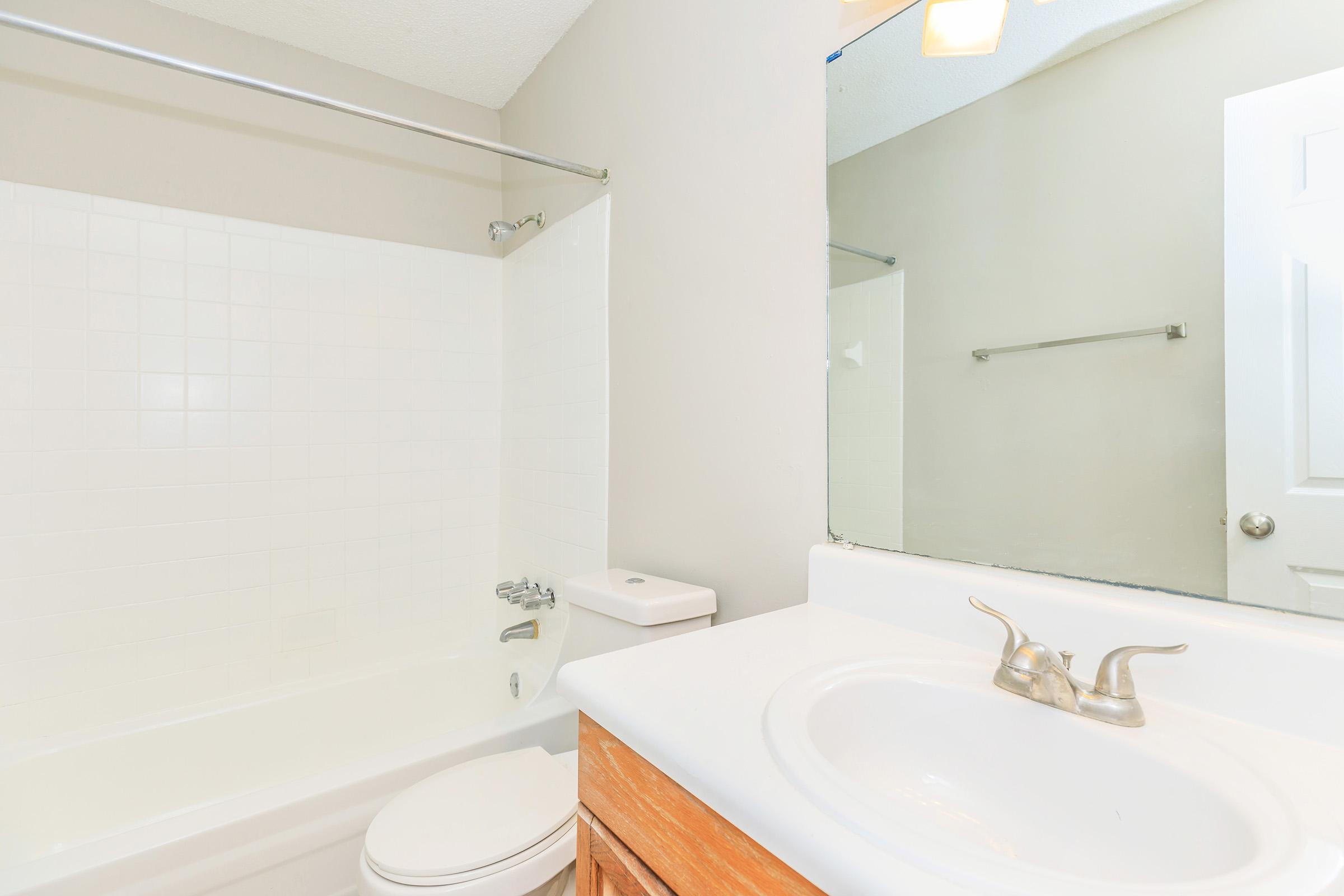 A clean and modern bathroom featuring a white bathtub with a shower fixture, a white sink with chrome faucets, and a large mirror above the sink. The walls are painted a light gray, and the space includes a towel bar and a door leading to another area.