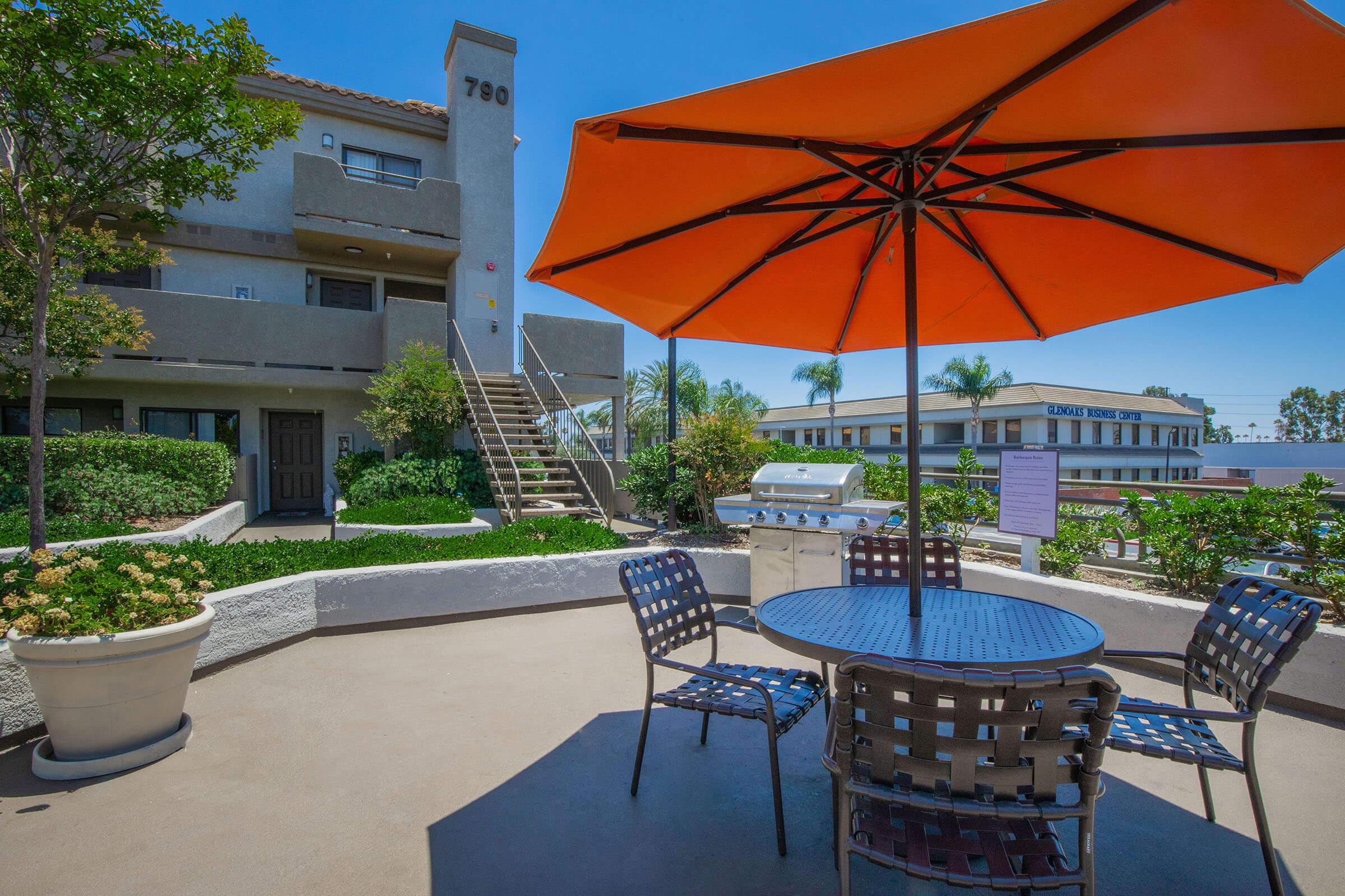 Outdoor patio area featuring a round table with four chairs, an orange umbrella for shade, and a barbecue grill nearby. The background includes a multi-story building with stairs and landscaped greenery, creating a welcoming atmosphere for relaxation and social gatherings.