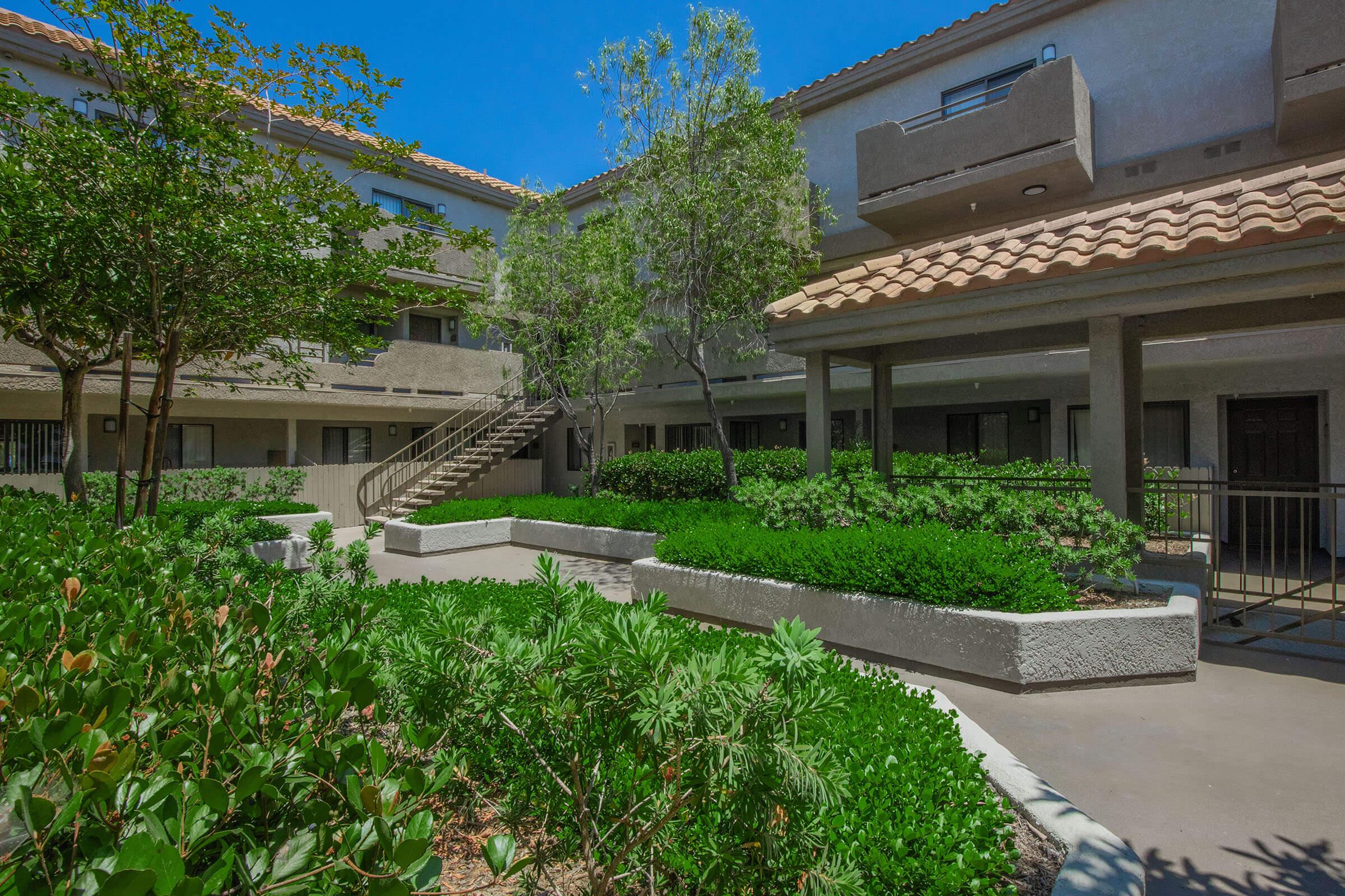 A view of a courtyard surrounded by low-rise buildings with a tiled roof. Lush greenery, including shrubs and small trees, adds to the serene atmosphere. A spiral staircase leads to upper levels, and pathways connect different areas of the courtyard under clear blue skies.