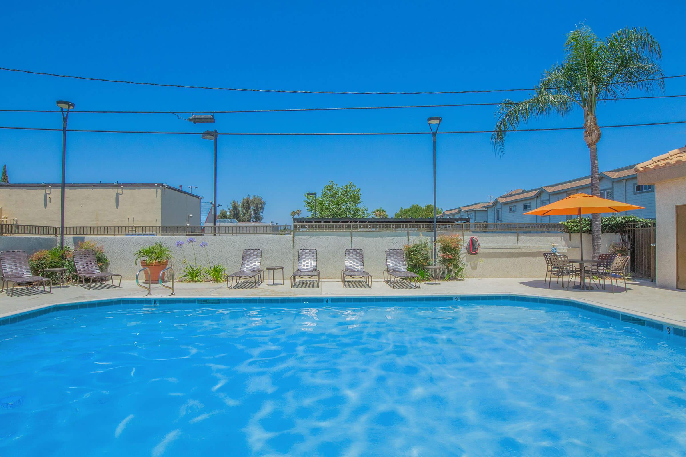 A swimming pool with clear blue water, surrounded by lounge chairs. There are palm trees and an orange umbrella providing shade. In the background, there are residential buildings and a clear blue sky. The area is well-lit with overhead lights.