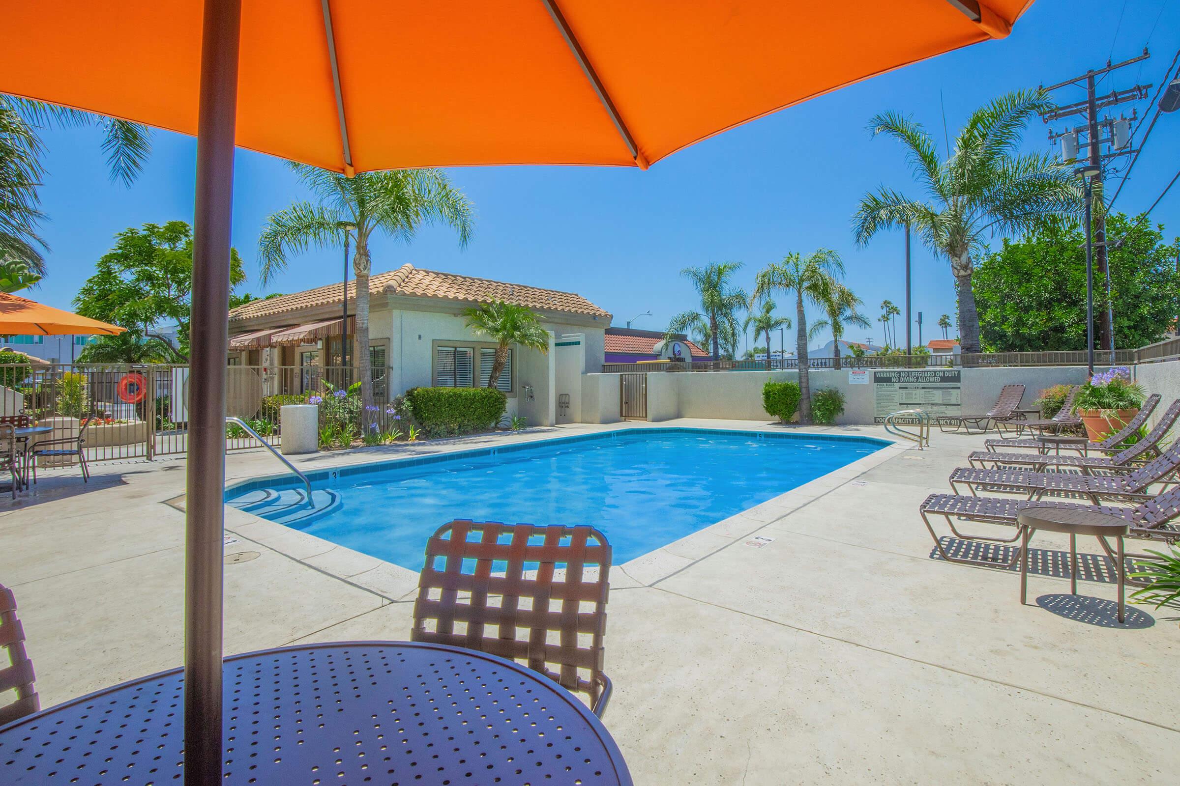 A clear swimming pool surrounded by lounge chairs and umbrellas, with palm trees and a sunny sky in the background. The area is well-maintained, featuring a building with a tiled roof. The scene conveys a relaxed, tropical atmosphere ideal for leisure and relaxation.