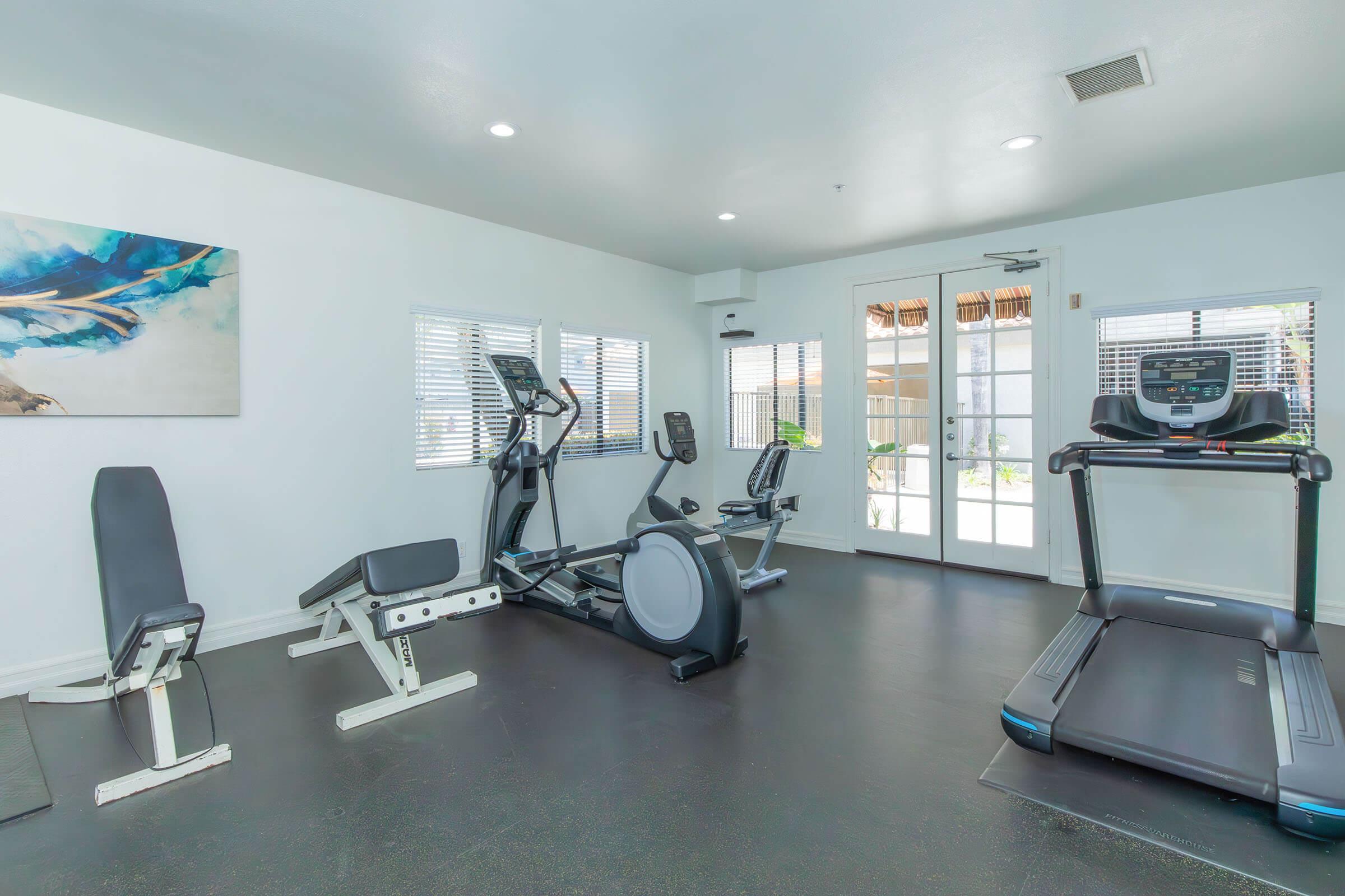 A modern fitness room featuring various exercise equipment including an elliptical machine, a stationary bike, a weight bench, and a treadmill. Bright, natural light enters through windows, and a painting hangs on the wall, creating a clean and inviting workout environment.