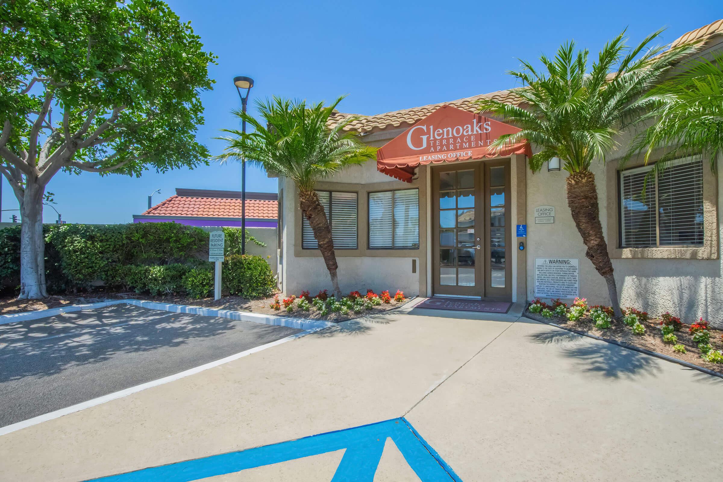 Exterior view of Glenoaks Treatment Center, featuring a welcoming entrance with a red awning and the center's name displayed. Surrounding greenery includes palm trees, and a well-maintained landscape with flowers. A parking area is visible, along with parking signs and clear blue skies.