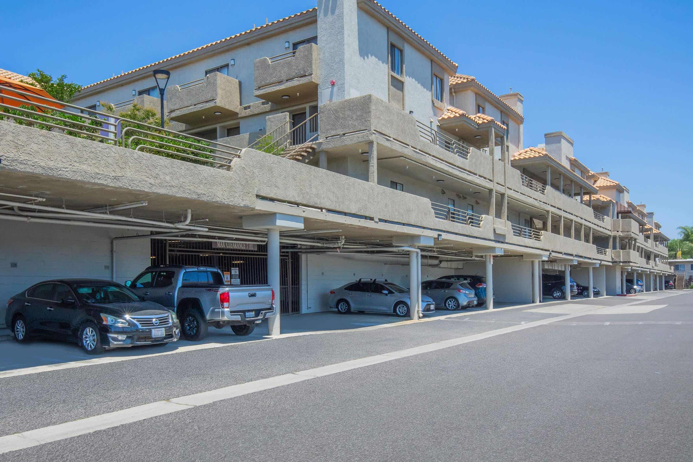 A row of multi-story residential buildings featuring covered parking spaces at the ground level. Several cars are parked under the structures, with a clear blue sky above and well-maintained surroundings. The scene conveys a modern residential area.