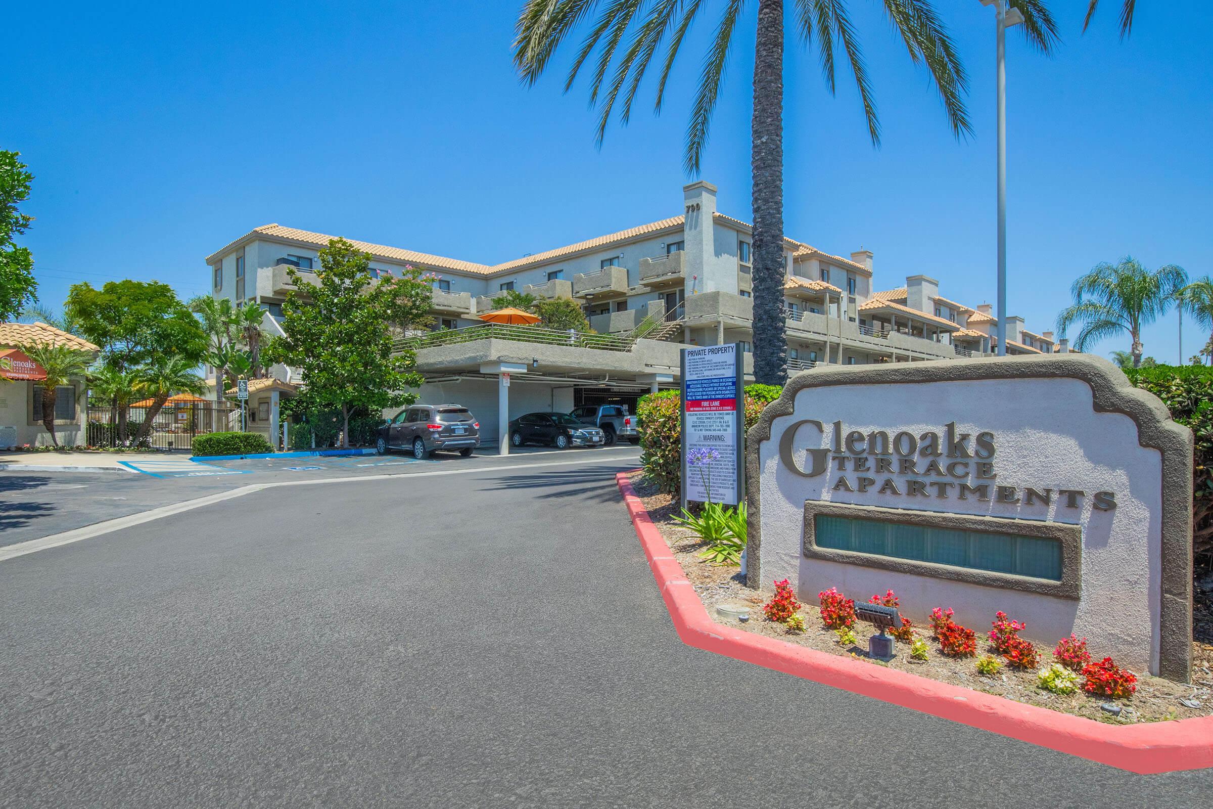 Sign for Glenoaks Terrace Apartments with landscaped entrance and palm trees. The parking area is visible with vehicles parked. The building features a Mediterranean architectural style under a clear blue sky. Red flowerbeds enhance the entrance.