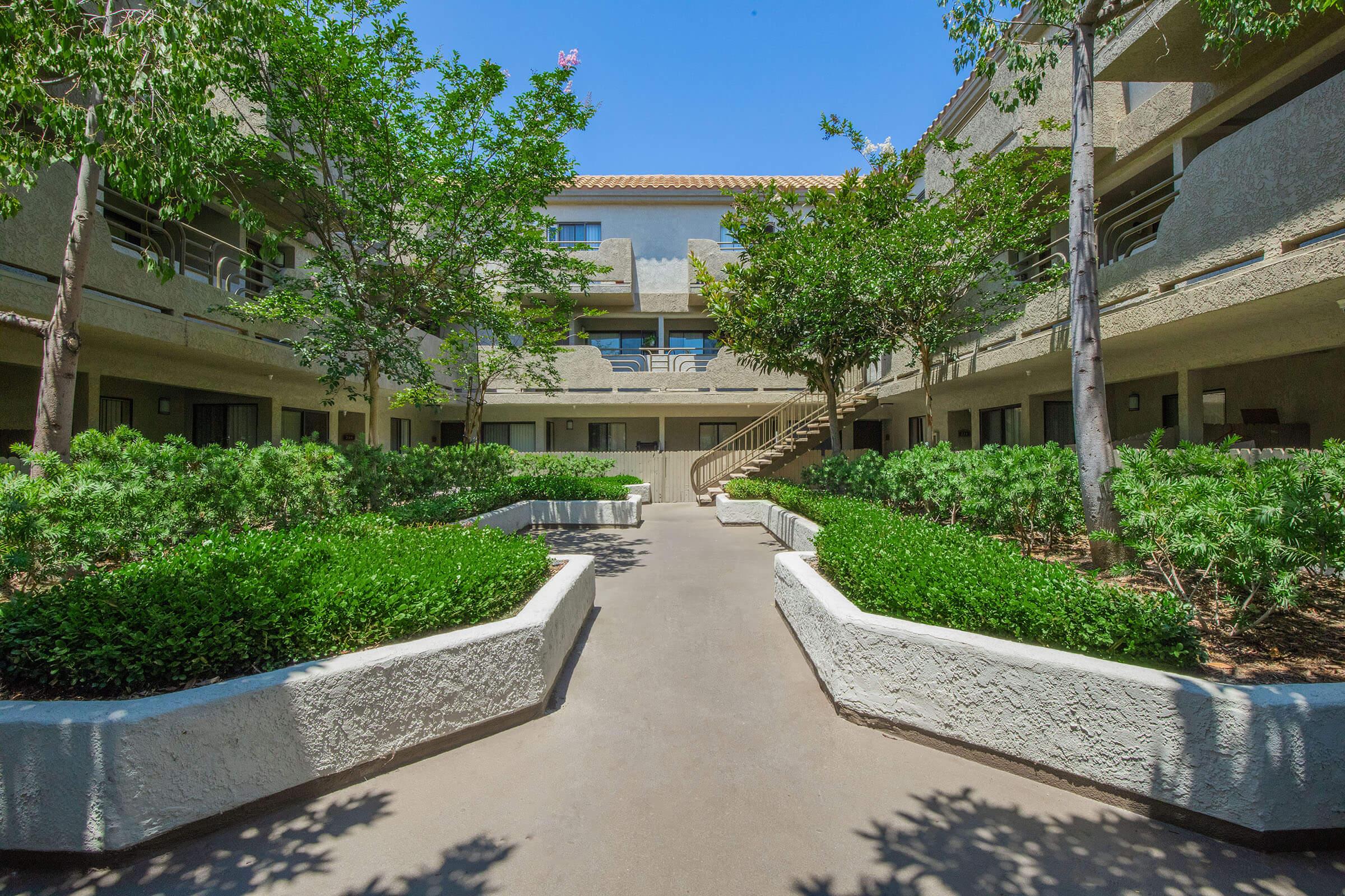A well-maintained courtyard of a multi-story building featuring green shrubs and trees. The pathway leads to the entrance of the building, with balconies visible on the upper level. The scene is bright and sunny, showcasing a pleasant outdoor space in a residential area.