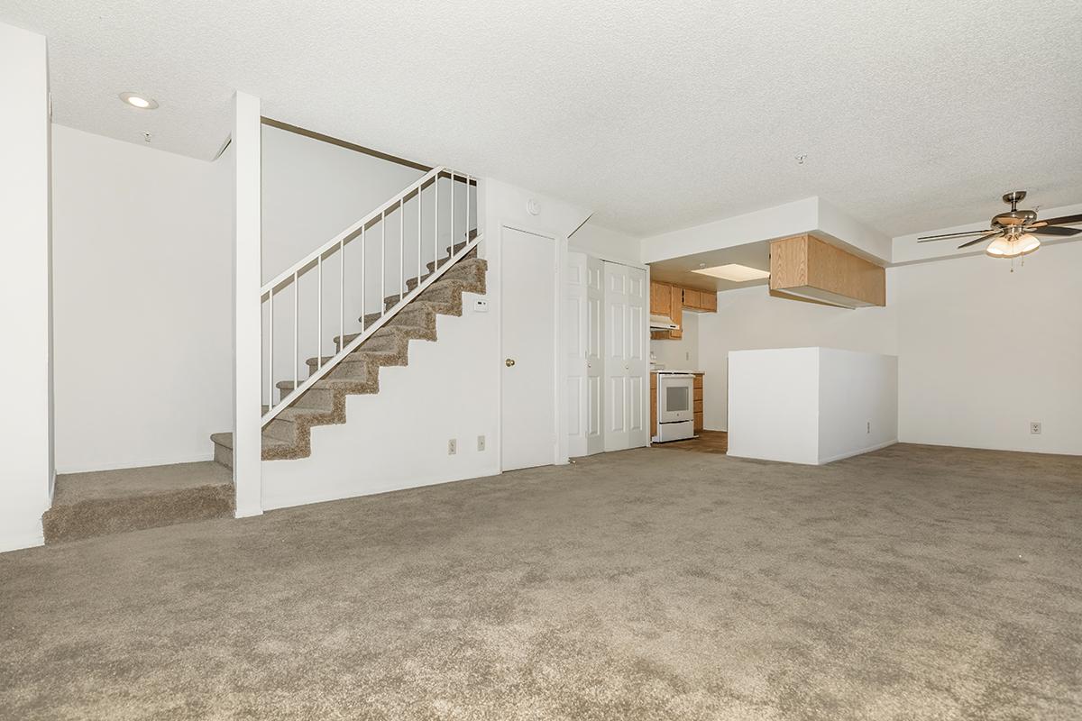 A spacious, carpeted living area with a staircase leading to an upper level. An open kitchen with wooden cabinetry is visible in the background. The walls are painted white, and there is a ceiling fan in the living space, creating a bright, inviting atmosphere.