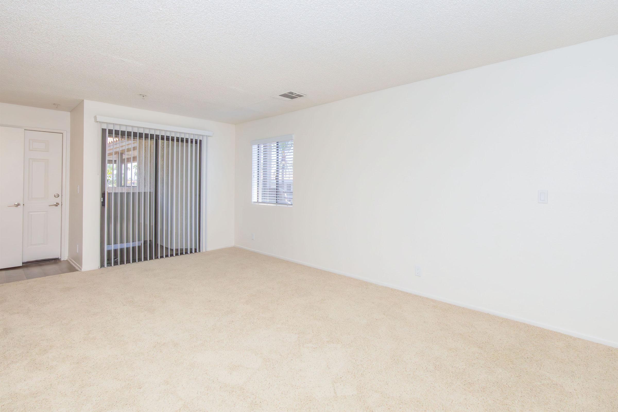 Empty room with beige carpet and white walls. There is a sliding glass door with vertical blinds on the left, leading to an outdoor area. A small window on the right lets in natural light, enhancing the spacious feel of the room. The overall atmosphere is bright and inviting, ready for furniture and decor.