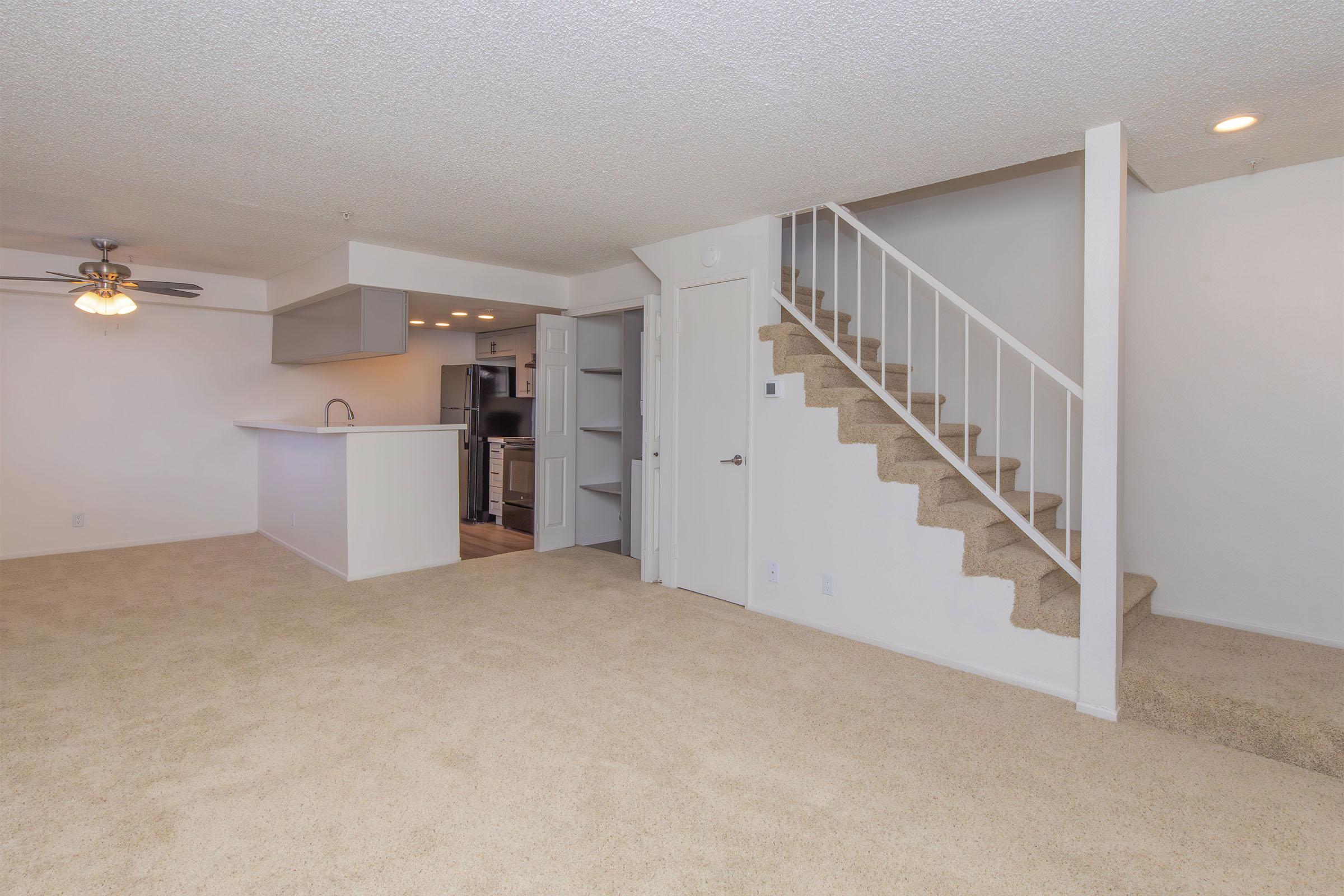 A spacious living area with beige carpet, featuring a ceiling fan. There is a staircase leading to an upper level, and an open kitchen space visible in the background. The walls are painted white, creating a bright and neutral atmosphere.