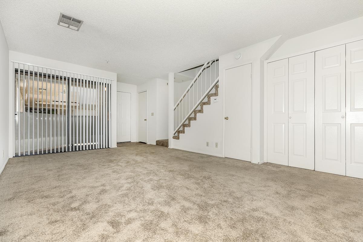 Empty living room area featuring beige carpet, a set of double doors leading to a hallway, a staircase against one wall, and a closet with sliding doors. The walls are painted white and there is a small window covered by vertical blinds.
