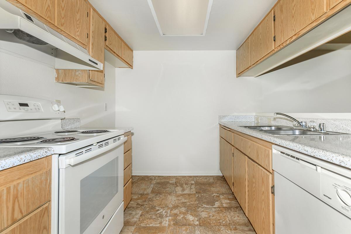 Modern kitchen featuring wooden cabinetry, a white oven and stovetop, a sink with a faucet, and a dishwasher. The countertops are light-colored, and the flooring is tiled. The overall space is bright and uncluttered with neutral walls, creating a spacious and functional cooking area.