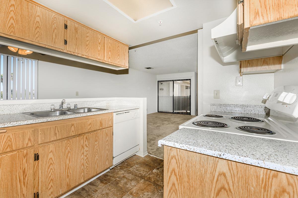A view of a kitchen featuring light wood cabinetry, a sink, a dishwasher, and a stove with an oven. The kitchen opens into a living space with neutral-colored walls and carpeting. Bright natural light is coming through the window blinds, providing an airy atmosphere.