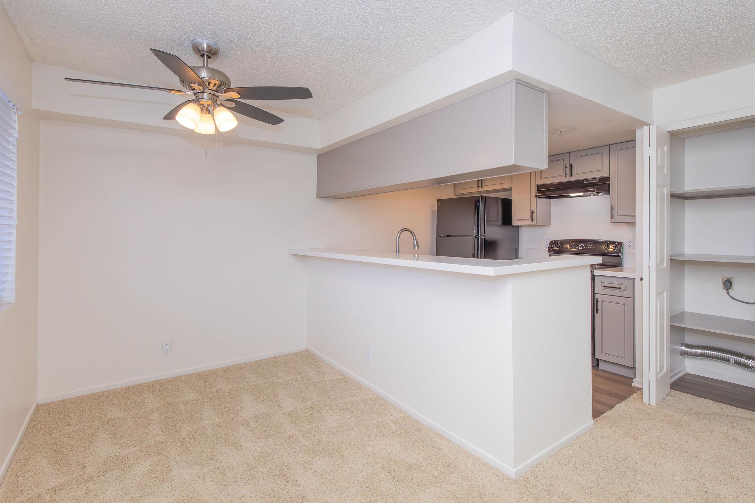 A well-lit, modern kitchen and living area featuring beige carpet flooring, a ceiling fan, and an open layout. The kitchen has light gray cabinetry, stainless steel appliances, and a breakfast bar. There is a window with light blinds, creating a bright atmosphere.