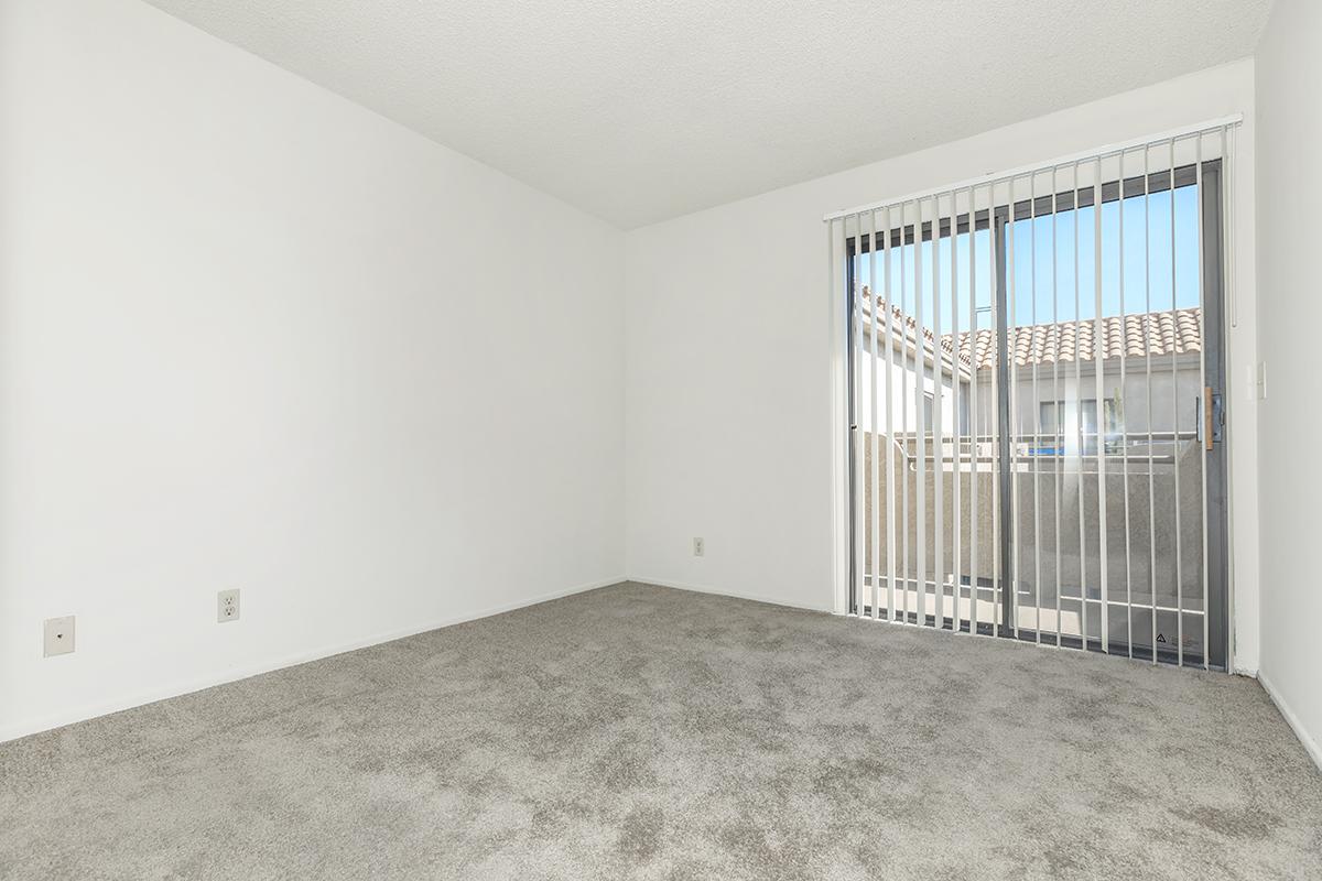 An empty room with light gray carpet and white walls. A sliding glass door with vertical blinds allows natural light to enter, leading to an exterior balcony or patio area. The space is unfurnished and spacious, creating a blank canvas for potential decoration.