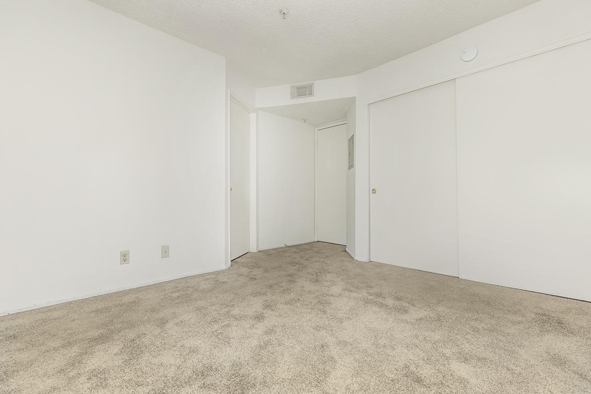 Empty room featuring light-colored walls and beige carpet. The space includes a doorway leading to another room, and two closed white closet doors. The overall ambiance is bright and minimalistic.