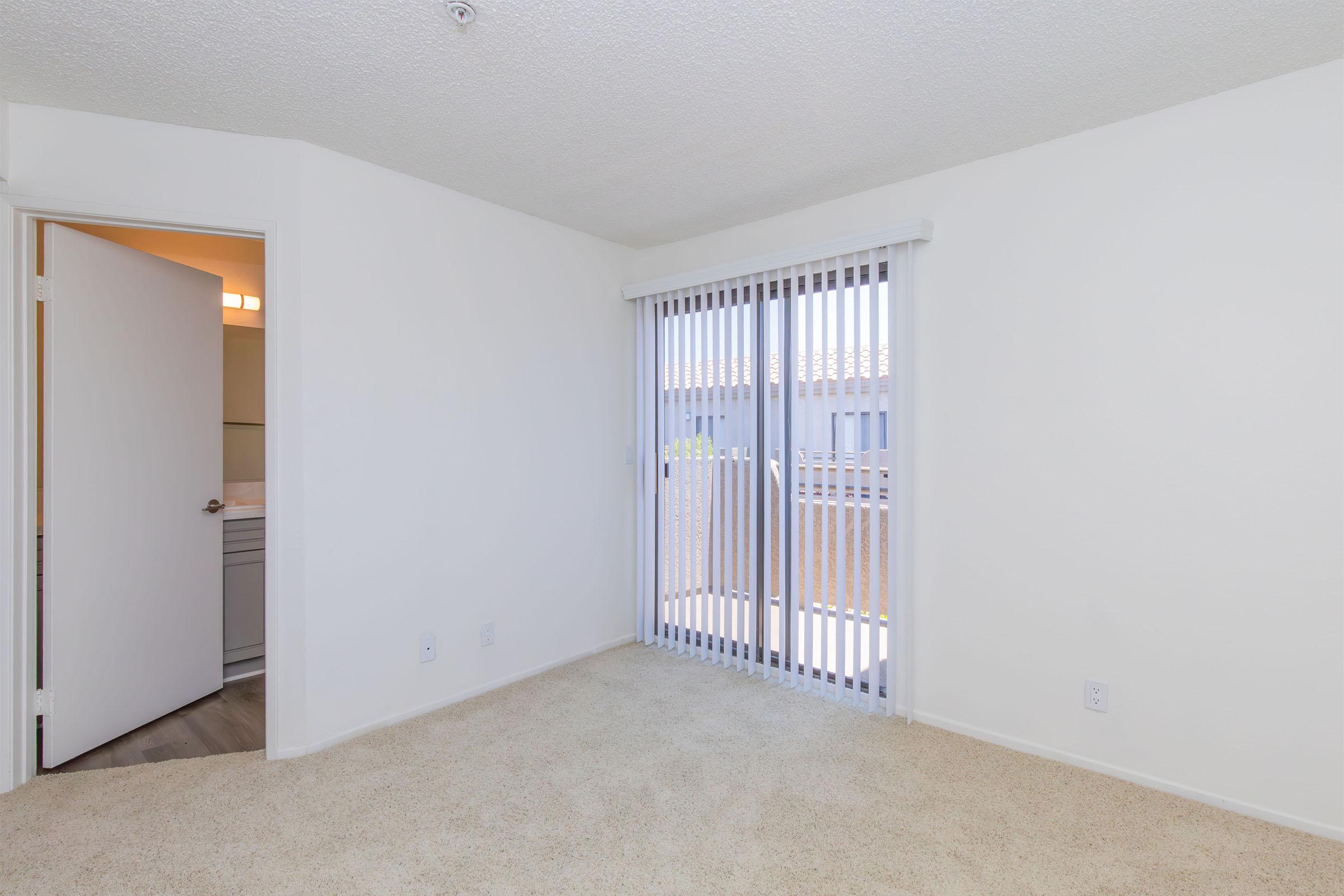 A bright, empty room with beige carpet and white walls. There is a sliding glass door with vertical blinds leading to an outdoor area. An open door on the left suggests access to another space. Natural light fills the room, creating an inviting atmosphere.