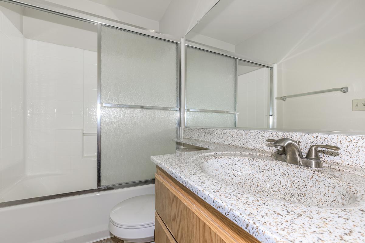 A modern bathroom featuring a glass shower enclosure, a bathtub, a sink with a speckled countertop, and wooden cabinetry. The space is well-lit and minimalistic, with a towel rack on the wall and clean, white tiles.