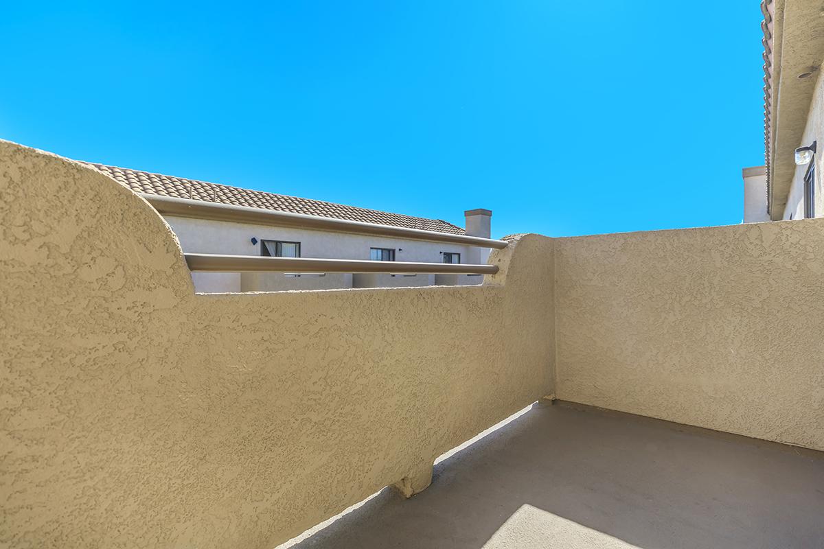 A light-colored balcony with a textured wall overlooking a clear blue sky. The balcony features a railing and is adjacent to a building with windows. The atmosphere conveys a bright and open outdoor space.