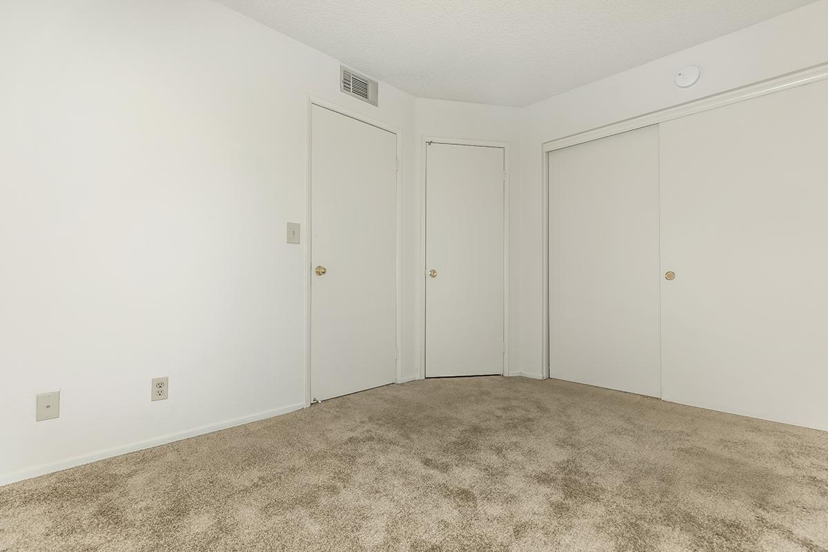 A simple, empty room with light beige carpet. It features white walls, with two closed doors: one leading to a closet and another a regular door. The ceiling has a light fixture, and there's a small vent on one wall. The overall atmosphere is neutral and uncluttered.