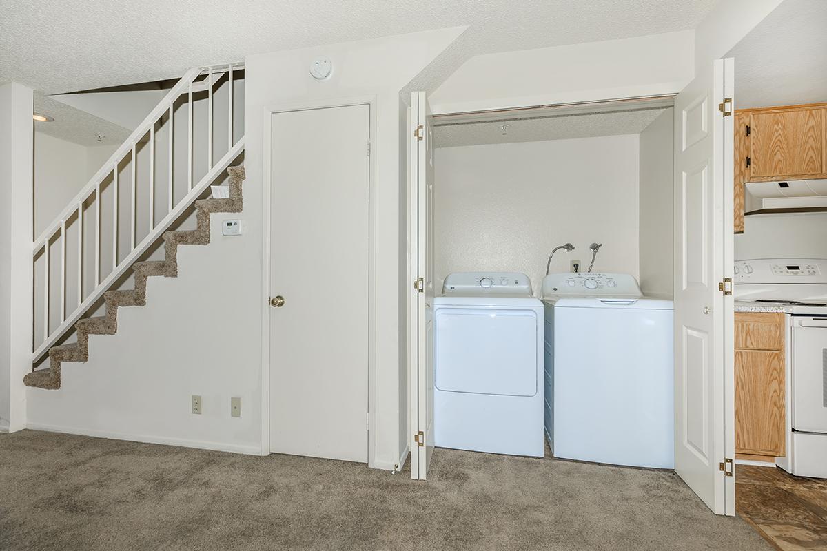 A laundry area featuring two white washing machines, located next to a staircase. The room has beige carpet and a light-colored wall, with a door leading to the laundry space partially open. A kitchen area is visible in the background, showcasing wooden cabinets and appliances.