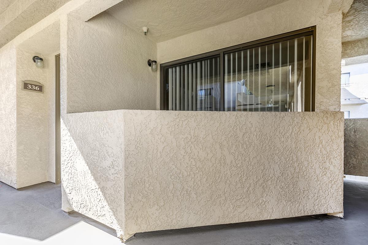 Exterior view of a residential building showing a textured wall and a window with vertical blinds. The wall is light-colored, and there is a view into a room through the window. A number plaque marked "336" is visible. The scene is well-lit, indicating a sunny day.