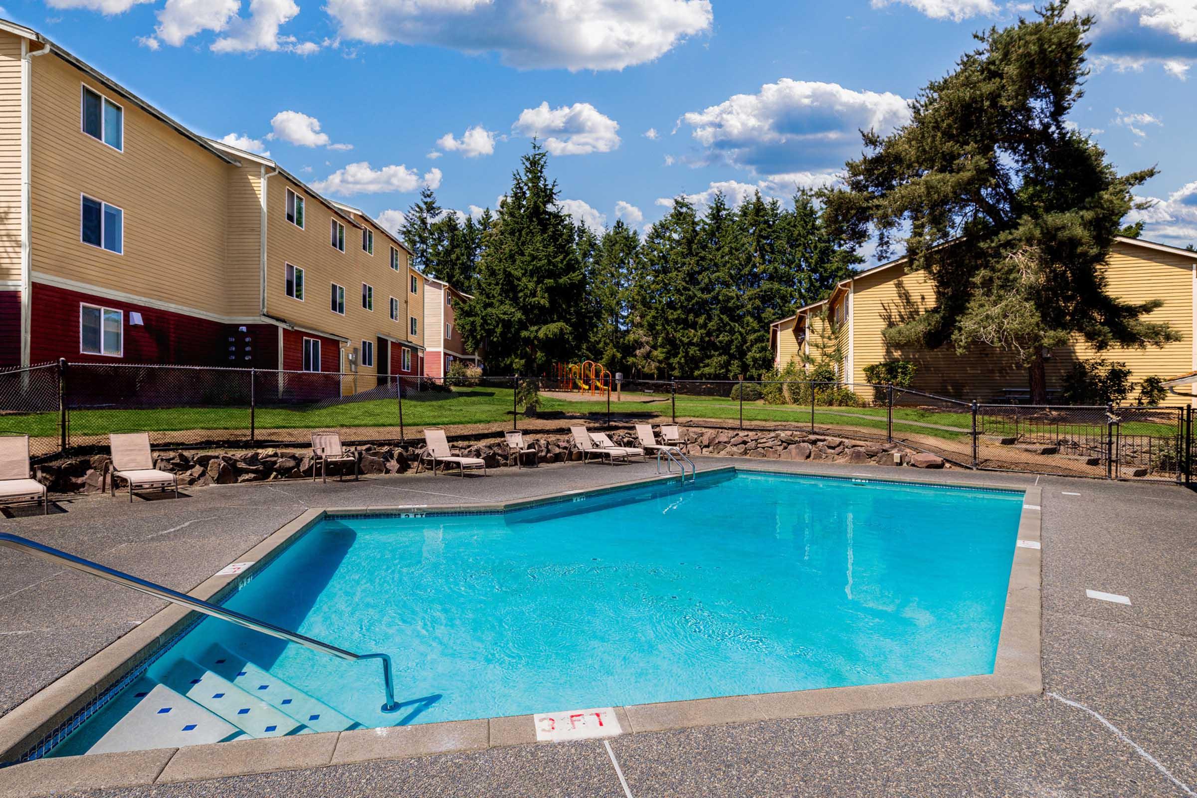 A clear blue swimming pool surrounded by lounge chairs, with a yellow building in the background. Tall trees and a playground are visible in the distance under a blue sky with scattered clouds. The pool has a "3 FT" depth marker at one end.
