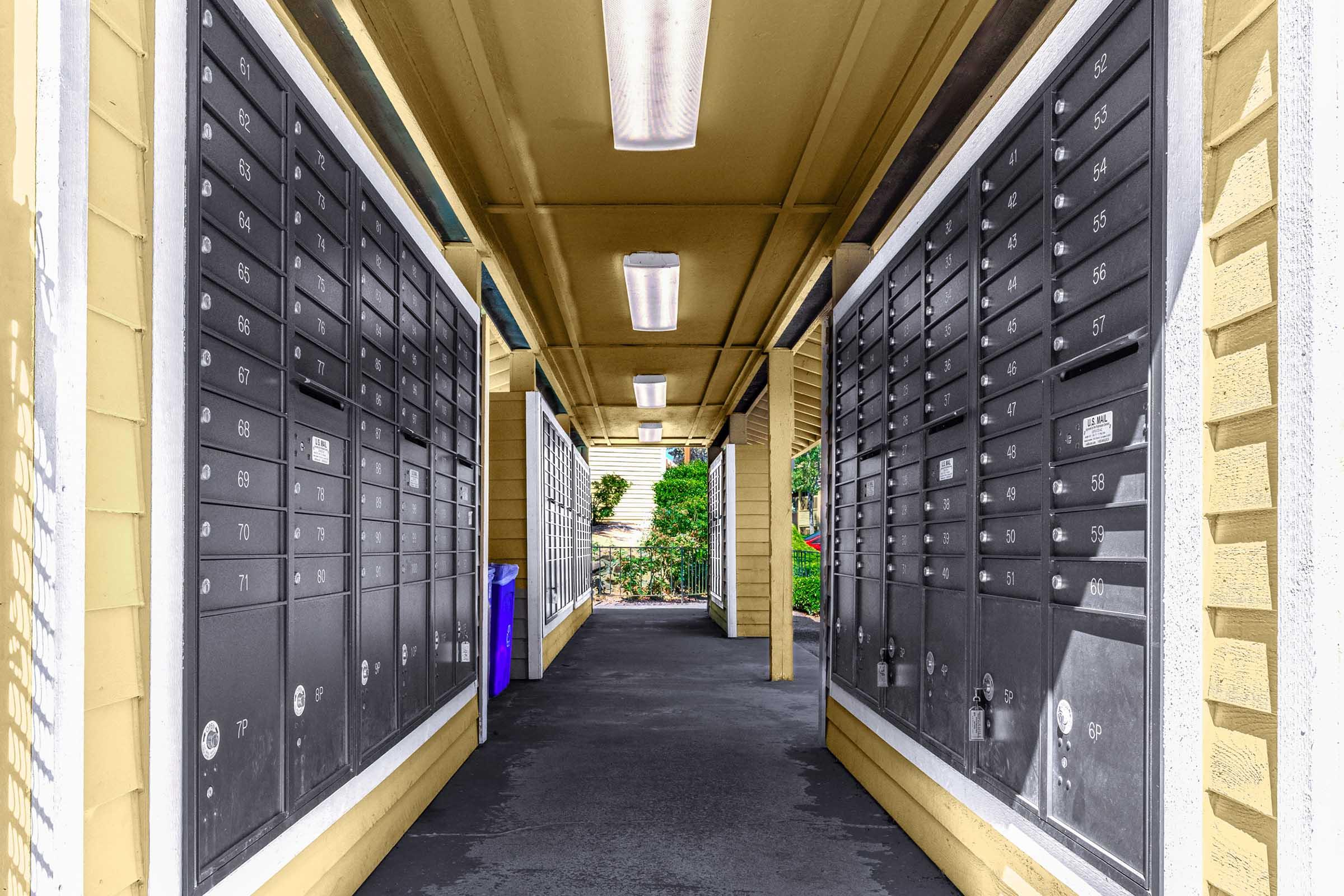 A row of black mailboxes lined on both sides of a covered walkway, illuminated by overhead lighting. The path is made of dark pavement, and there are hints of greenery visible at the end of the corridor. Some of the mailboxes have numbers displayed on them.