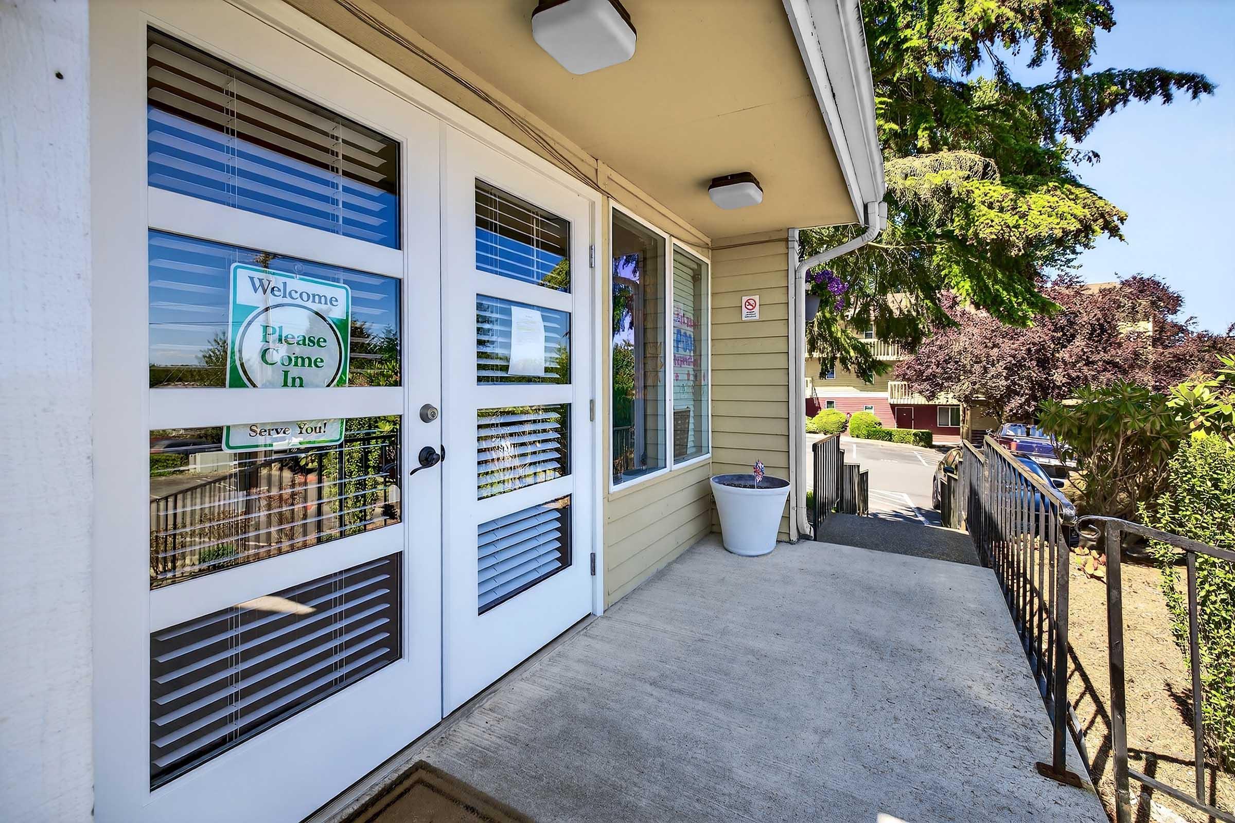 A welcoming entrance with double doors featuring a "Welcome" sign. The pathway leads to a well-maintained exterior with potted plants and greenery on either side. Natural light enhances the inviting atmosphere of the entryway.