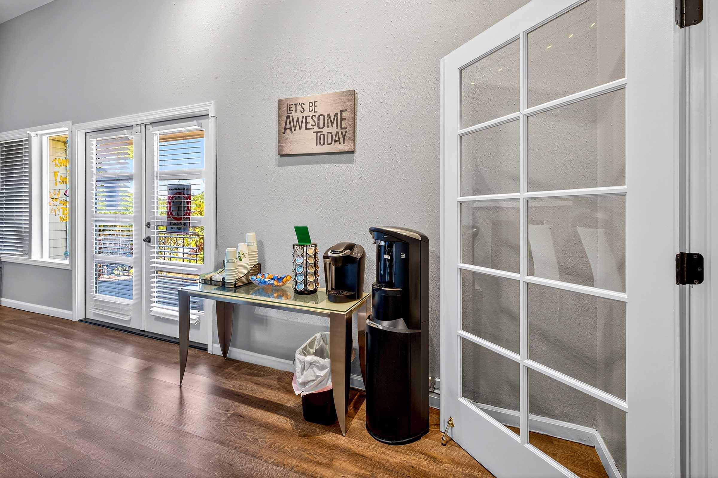 A well-lit room featuring a table with refreshments, including cups and snacks, next to a coffee machine. There are large windows with blinds, allowing natural light to enter. A motivational sign on the wall reads, "LET'S BE AWESOME TODAY." A white door leads to another area. The flooring is wooden.