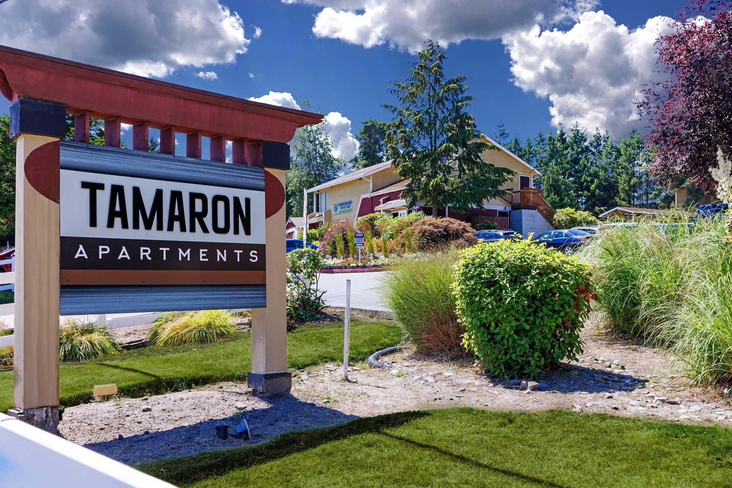 Signage for Tamaron Apartments, featuring a large, clearly visible name on the sign. The scene includes well-manicured landscaping with shrubs and grass, and in the background, the apartment building is partially visible under a blue sky with fluffy white clouds.