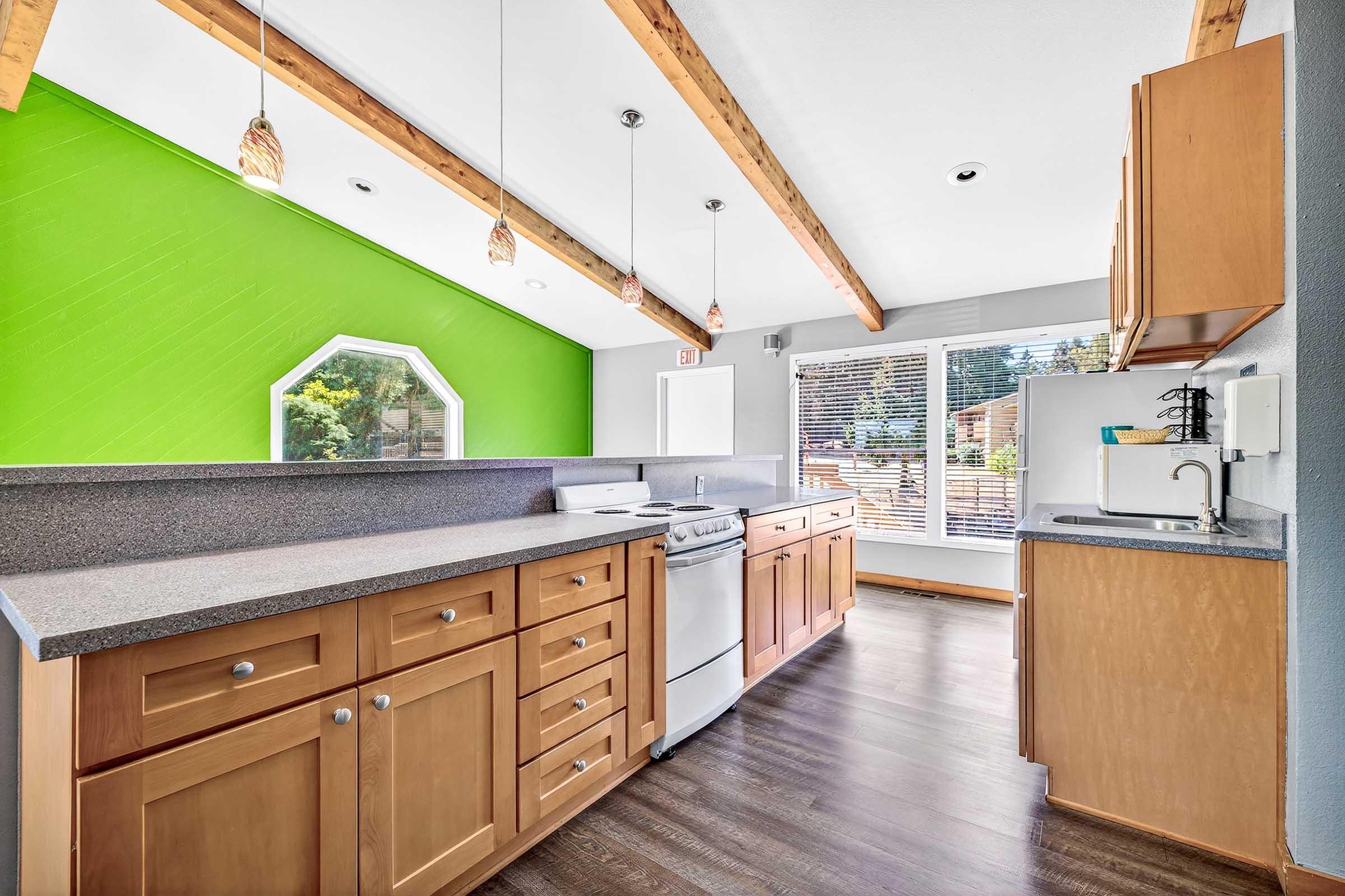 Modern kitchen featuring wooden cabinetry, gray countertops, and stainless steel appliances. Bright green accent wall complements the open space, with natural light streaming through large windows. Exposed wooden beams add character to the ceiling, creating a warm and inviting atmosphere.