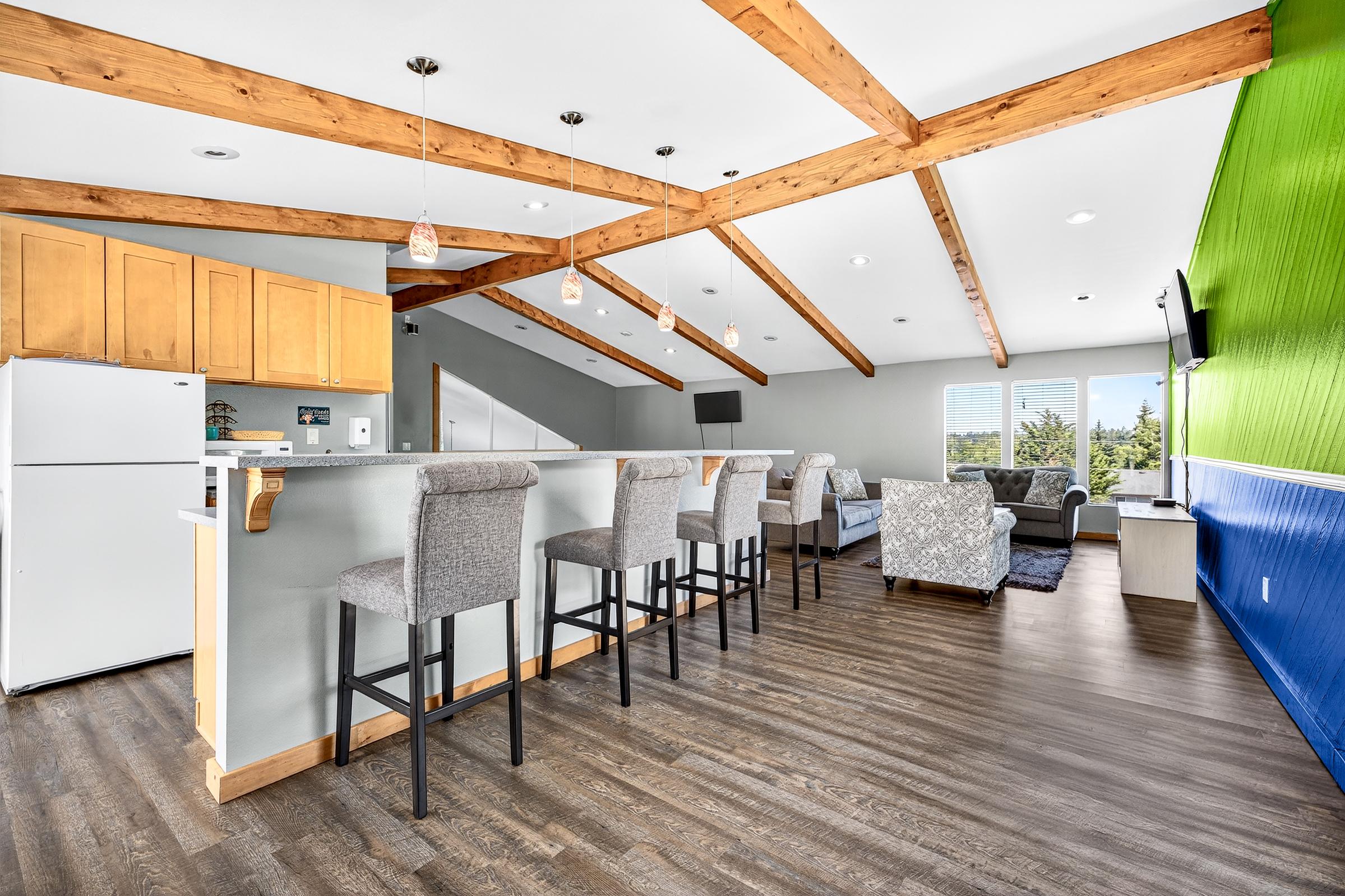 A modern living space featuring a kitchen bar with stools, wood beam ceilings, and a cozy seating area. The walls are painted in a vibrant green and neutral tones. A refrigerator is visible, along with a flat-screen TV, creating a bright and inviting atmosphere.