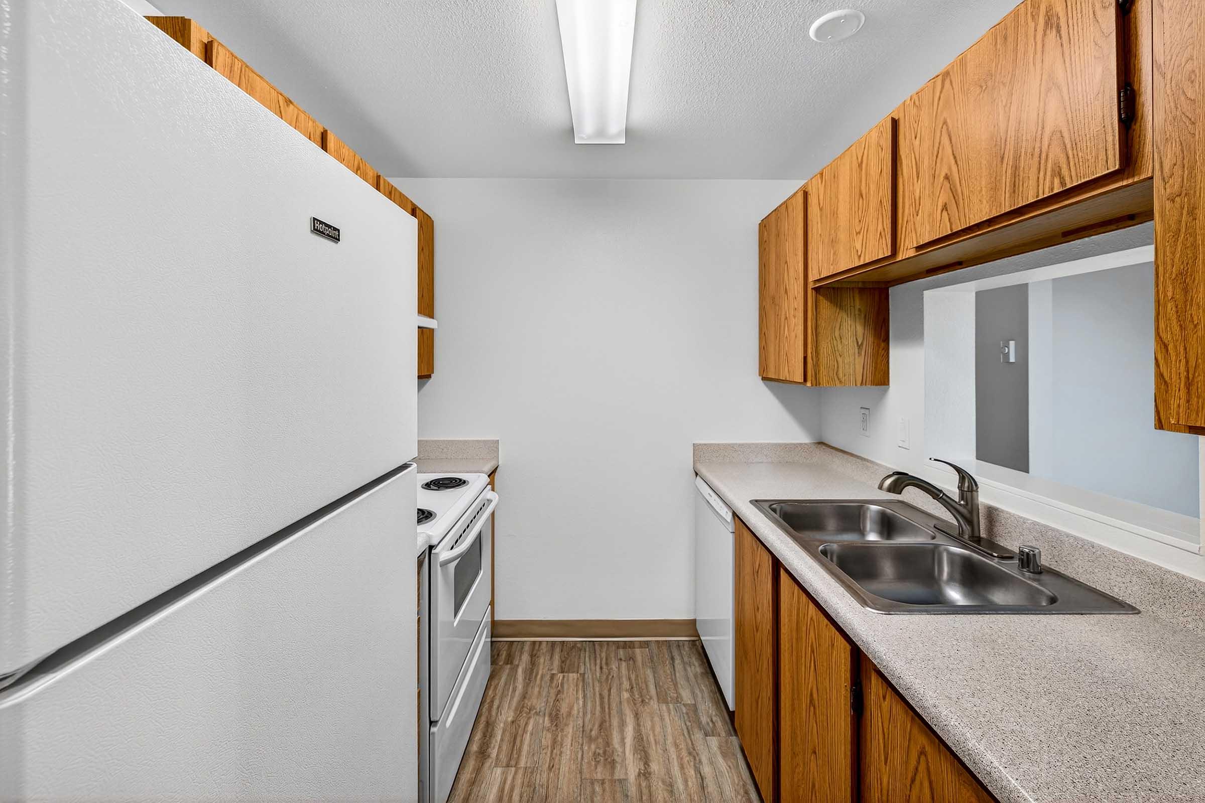 A small kitchen with wooden cabinets, a white refrigerator, a white stove, and a double stainless steel sink. The countertops are light-colored, and the floor features wood-like laminate. There is a bright overhead light providing illumination.
