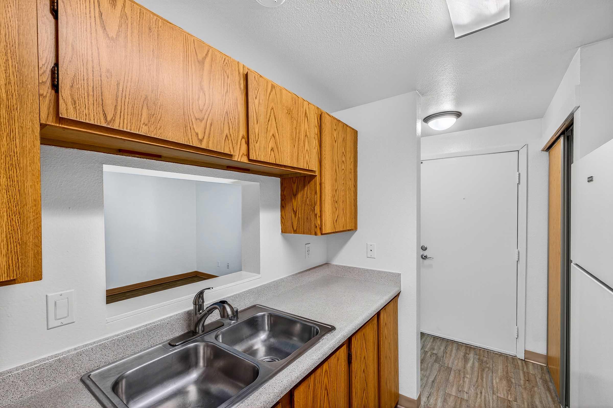 A modern kitchen featuring wooden cabinets, a countertop with a double sink, and an open layout that connects to a nearby wall. A doorway leads to an entry area with a light-colored wall and flooring, along with a white refrigerator visible in the corner.