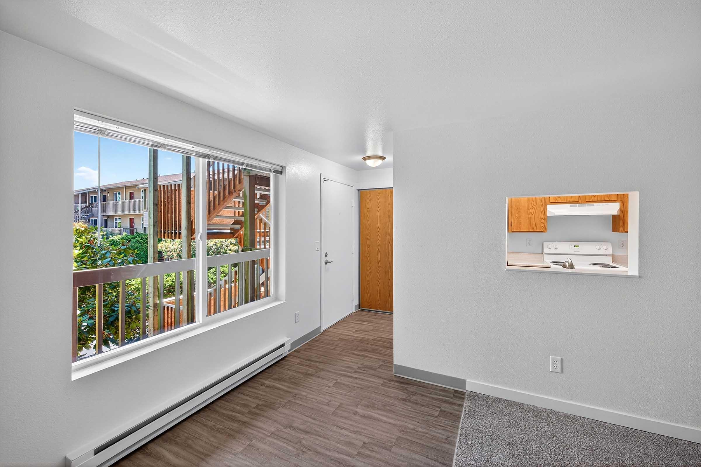 Bright and inviting interior of an apartment featuring a large window with a view of greenery, a door leading outside, and a partial glimpse of the kitchen. The walls are painted white, floors are a mix of carpet and laminate, and there is a small, open wall cutout connecting to the kitchen area.