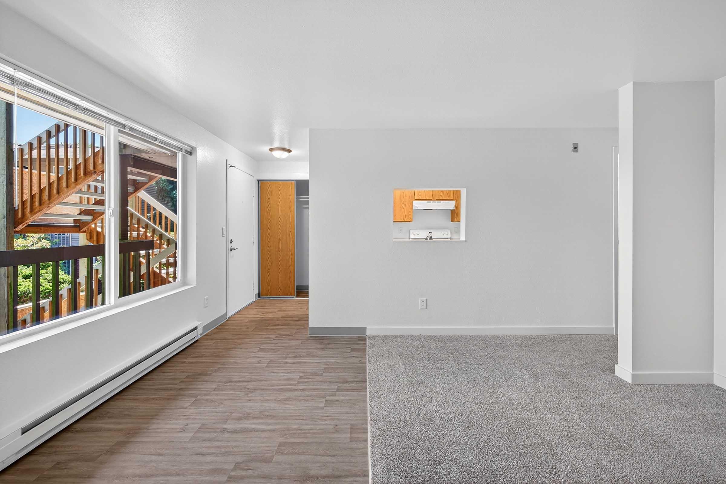 Bright and spacious interior of an apartment featuring a large window with natural light, a carpeted area, and a wood-style floor. There is a small entryway with a closet and a mirror on the wall. The walls are painted white, and a wooden staircase is visible through the window, indicating access to multiple levels.