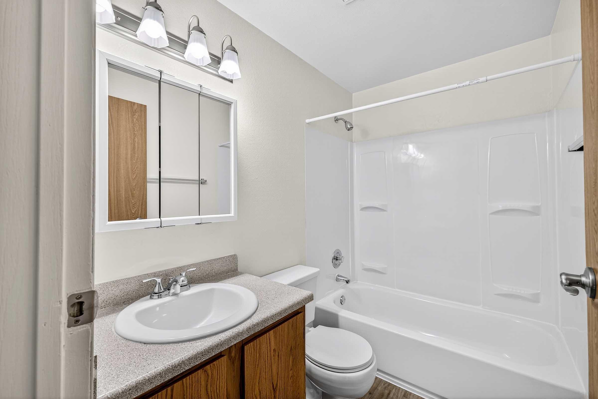 A clean and well-lit bathroom featuring a white bathtub and shower, a sink with a wooden cabinet below, a mirror above the sink, and a toilet. The walls are painted in a light color, and the floor has a wooden appearance. The bathroom is organized and tidy, with modern light fixtures.