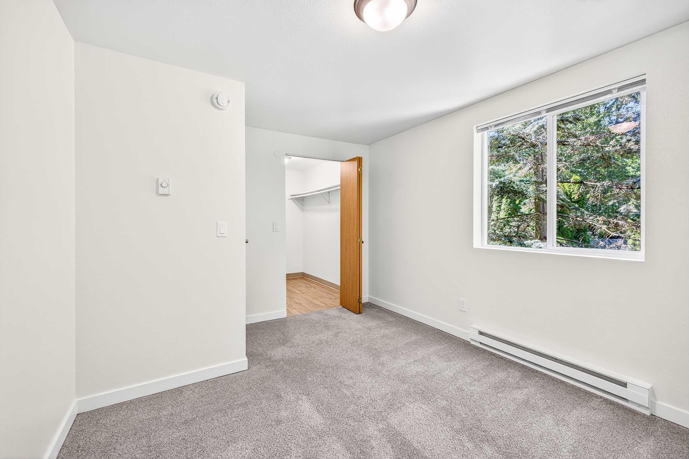 A bright, empty room featuring light-colored walls and a large window overlooking greenery. The floor is carpeted in a neutral tone, and there is a closet with open shelving on one side. The room is well-lit, creating a welcoming atmosphere.