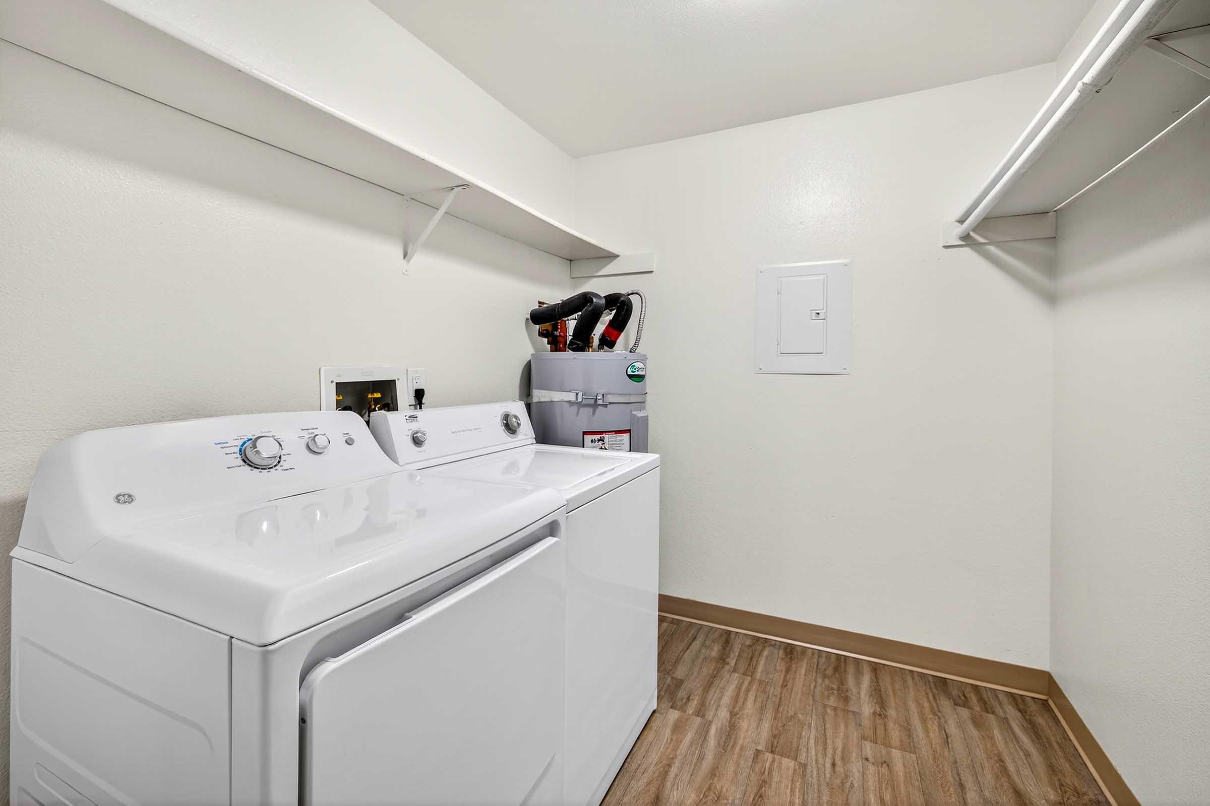 A clean and bright laundry room featuring a stacked washer and dryer, with a small shelf above the appliances. There's a wall-mounted electrical panel and a hanging rod for clothes. The flooring is a light wood design, and the walls are painted in a light color, creating an open and spacious feel.