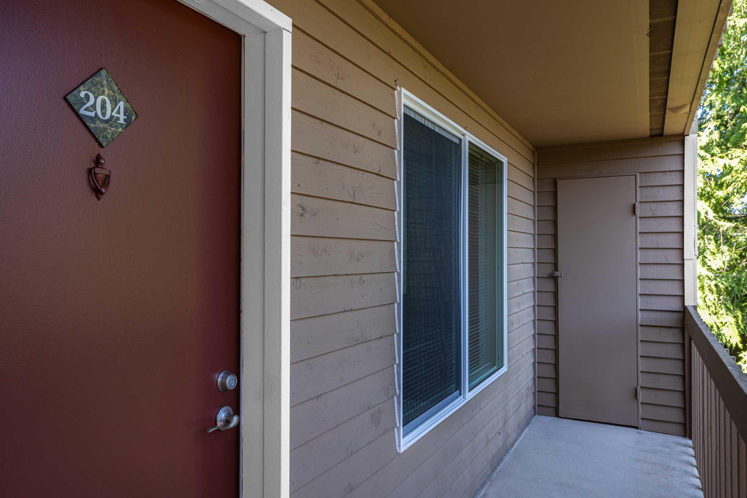 A close-up view of an apartment entrance featuring a brown door with the number "204" displayed on a diamond-shaped plaque. There is a window beside the door, and a second door can be seen to the right. The exterior walls are beige, and the balcony is covered with a light gray surface.