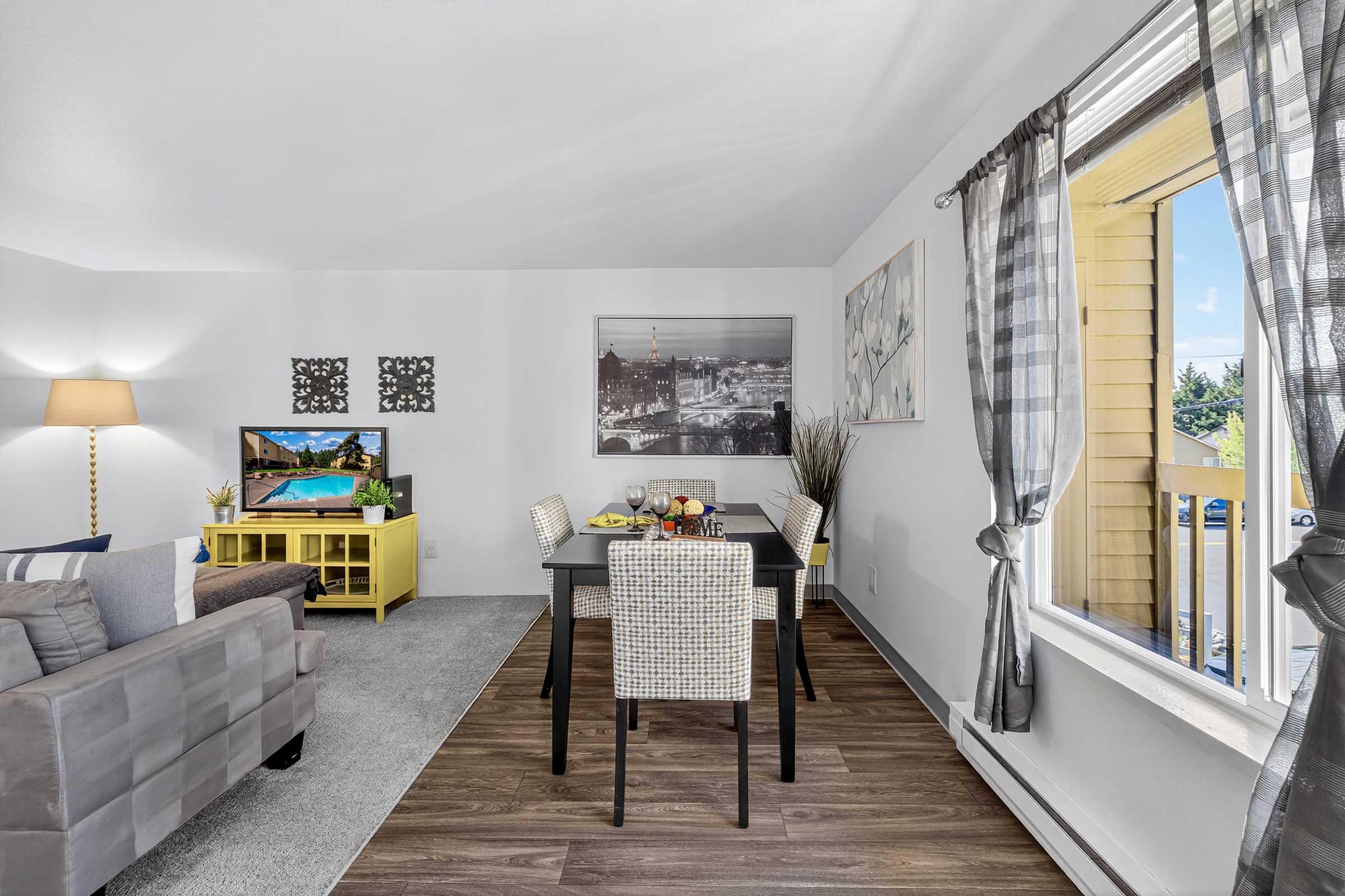 Living room and dining area featuring a cozy gray sofa, a yellow media console with a TV, and a dining table set for two. Large windows with patterned curtains let in natural light. Tasteful wall art and a view of a balcony add charm to the modern decor.