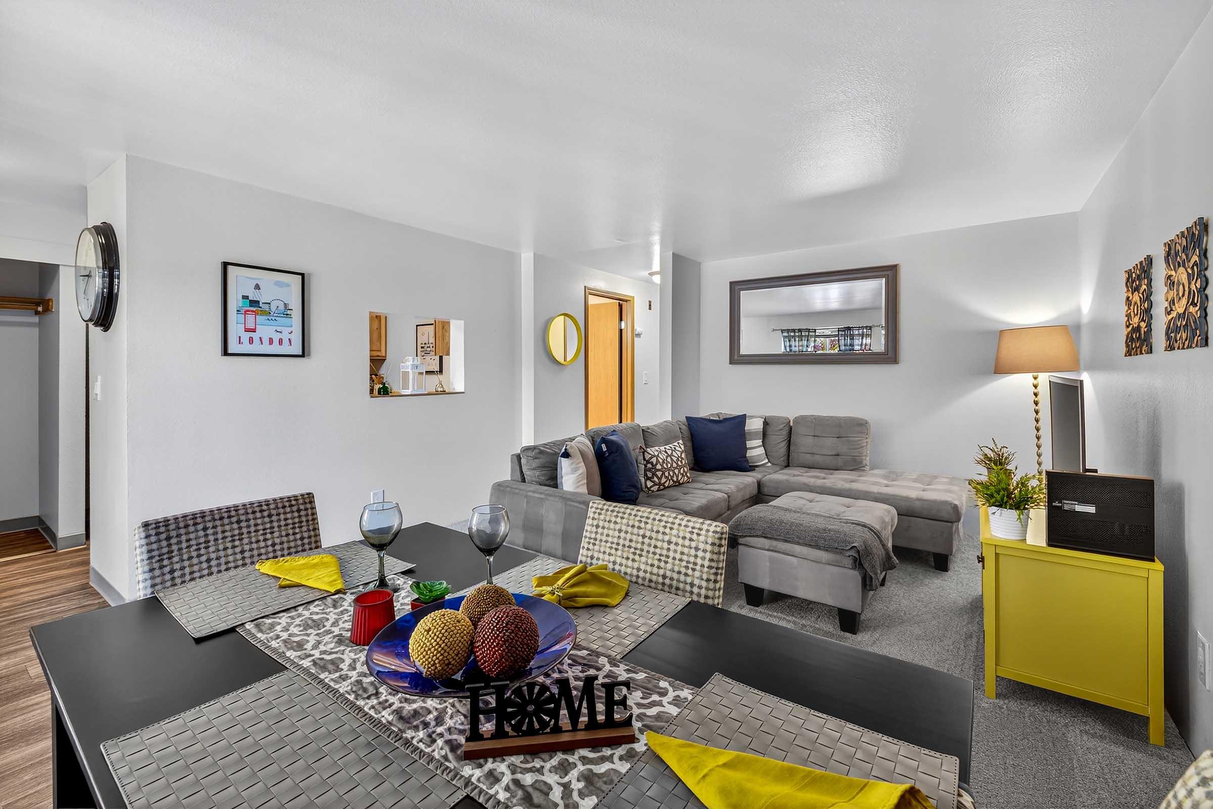 A cozy living room featuring a sectional sofa, decorative pillows, and a coffee table. A dining table with yellow napkins and a centerpiece of decorative balls is set nearby. The room has light gray walls, a mirror, and artwork, creating a warm and welcoming atmosphere.